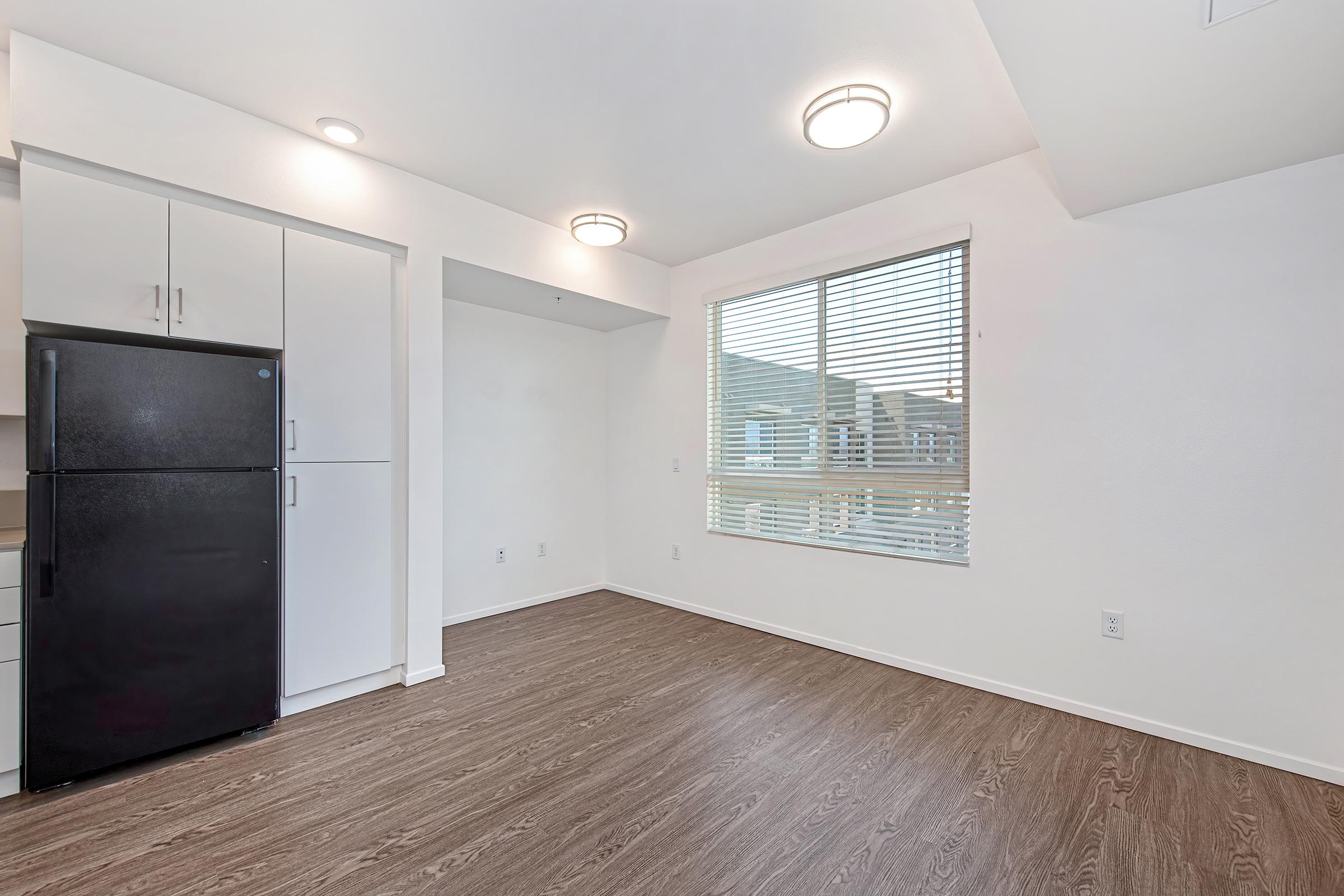A modern, empty kitchen space featuring a black refrigerator, white cabinetry, and a large window with blinds. The room has laminate wood flooring and recessed lighting, creating a bright and open atmosphere.