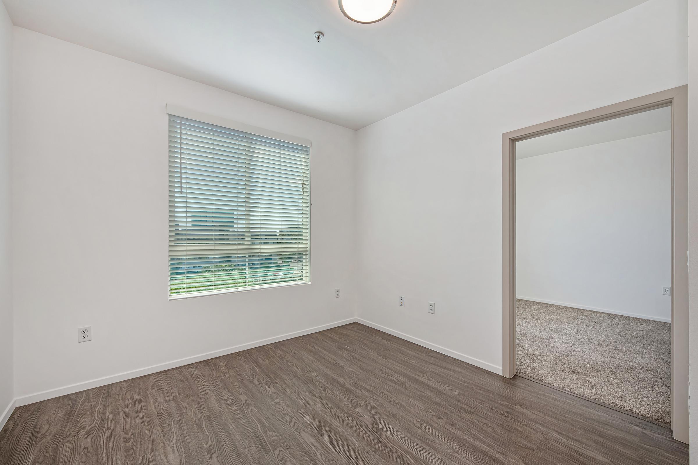 Empty room with light brown laminate flooring, a window with horizontal blinds letting in natural light, and a neutral-colored wall. A doorway leads to another space, suggesting a connected layout. The overall atmosphere is bright and clean, suitable for various purposes.
