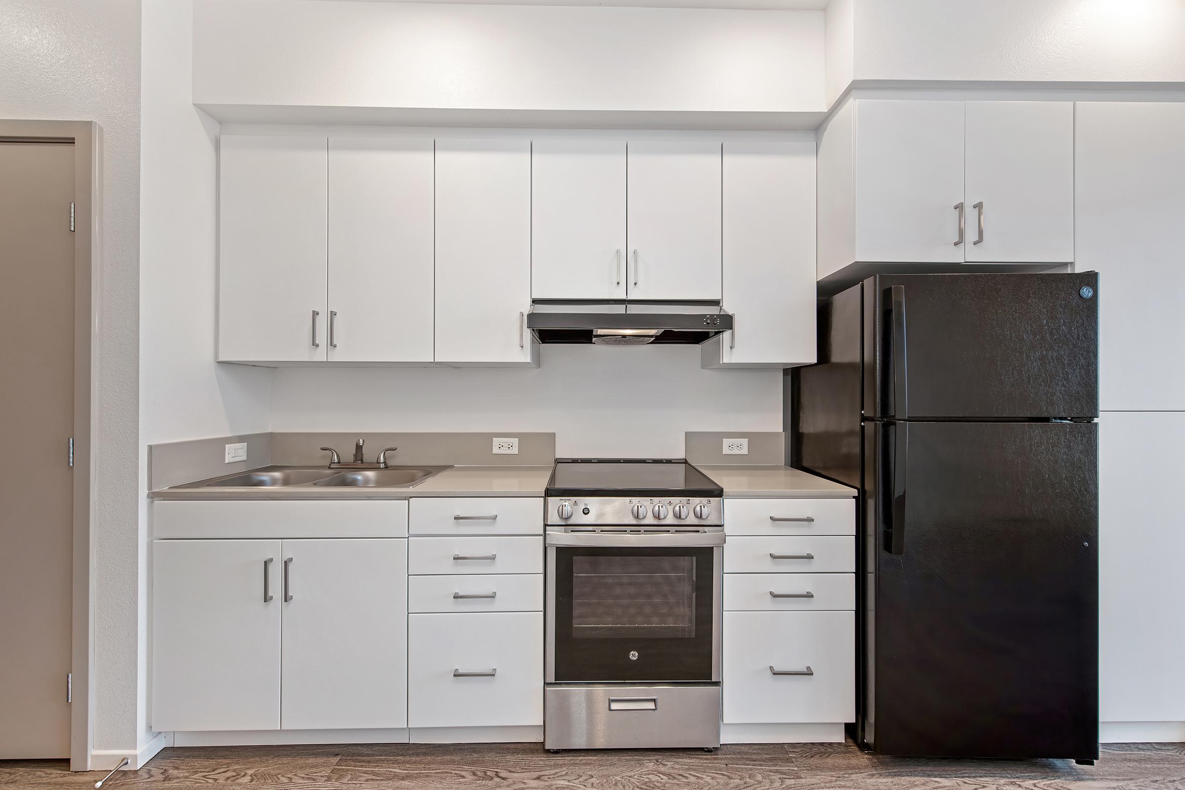 Modern kitchen featuring white cabinets, a black refrigerator, a stainless steel stove and oven combination, and a sink. The countertop is light-colored, and the flooring is wooden, creating a contemporary look.