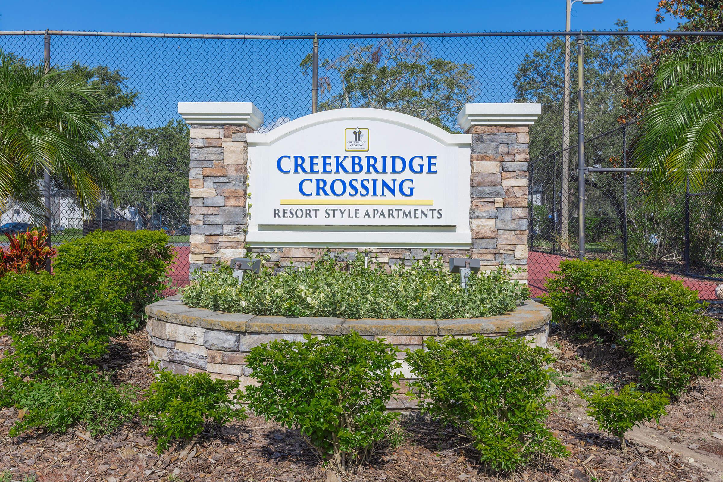 Sign for Creekbridge Crossing with the text "Creekbridge Crossing Resort Style Apartments." The sign is set in a landscaped area with green plants and a stone border, against a backdrop of trees and a clear blue sky.