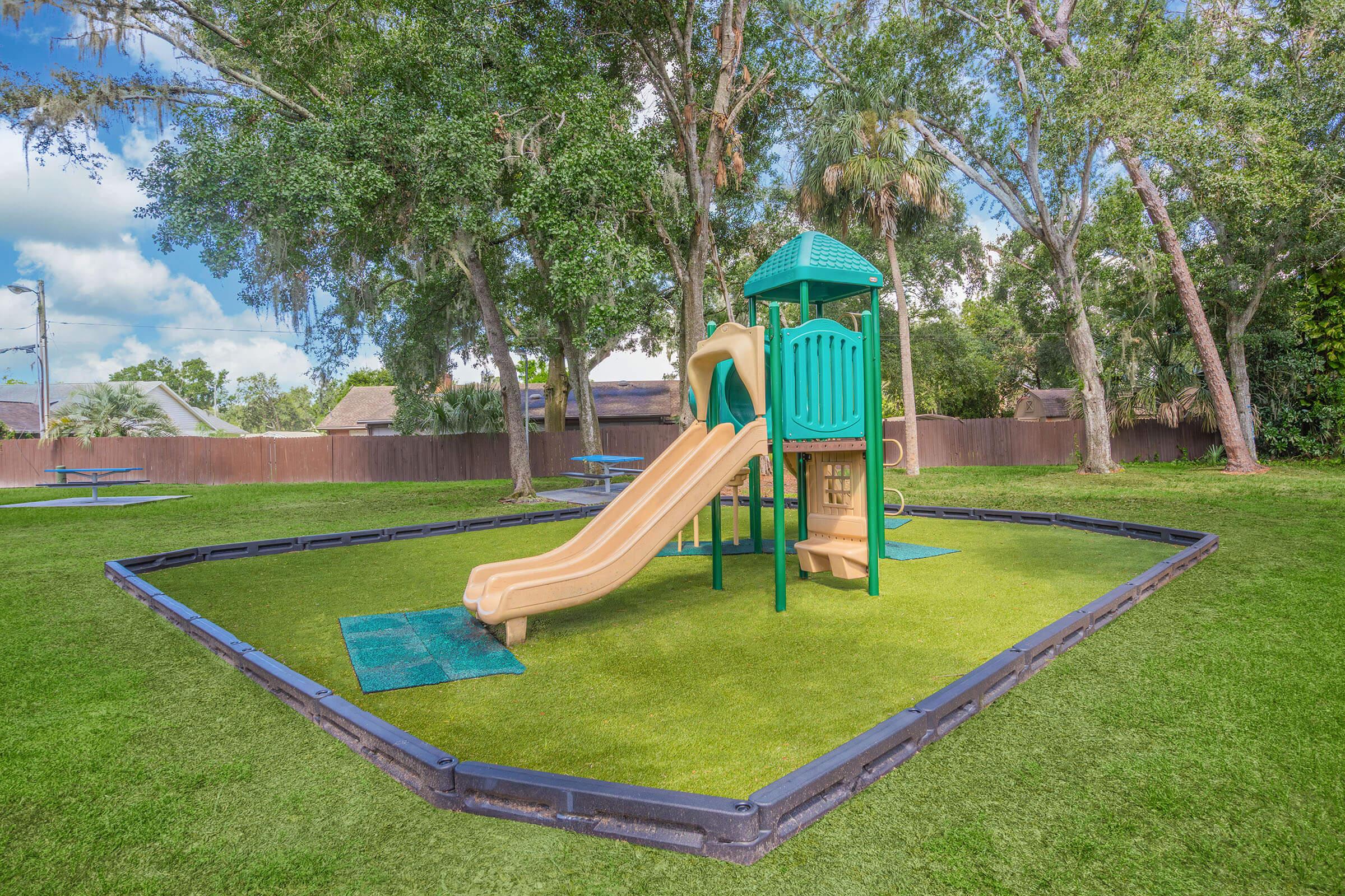 A colorful playground featuring a green and tan slide and climbing structure, set on a grassy area surrounded by trees. Picnic tables are visible in the background, and the sky is partially cloudy.