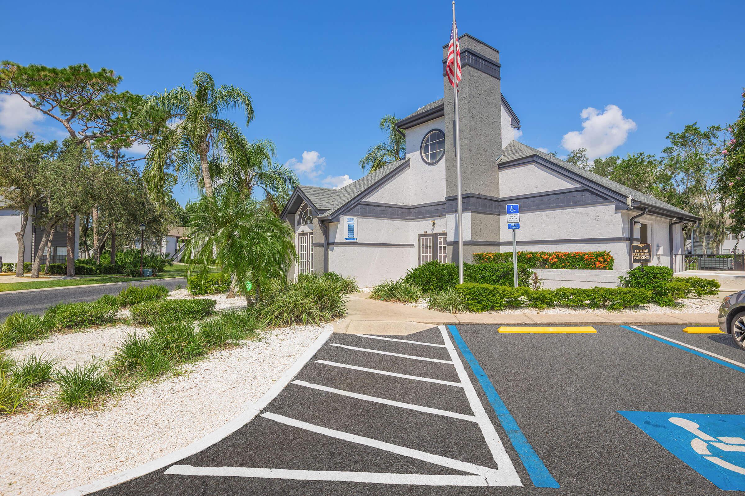 A modern building with a gray exterior and a pointed roof, surrounded by palm trees and manicured landscaping. A parking area with clear blue skies overhead. An American flag is flying near the entrance, which has a welcoming appearance. The scene conveys a vibrant and pleasant atmosphere.