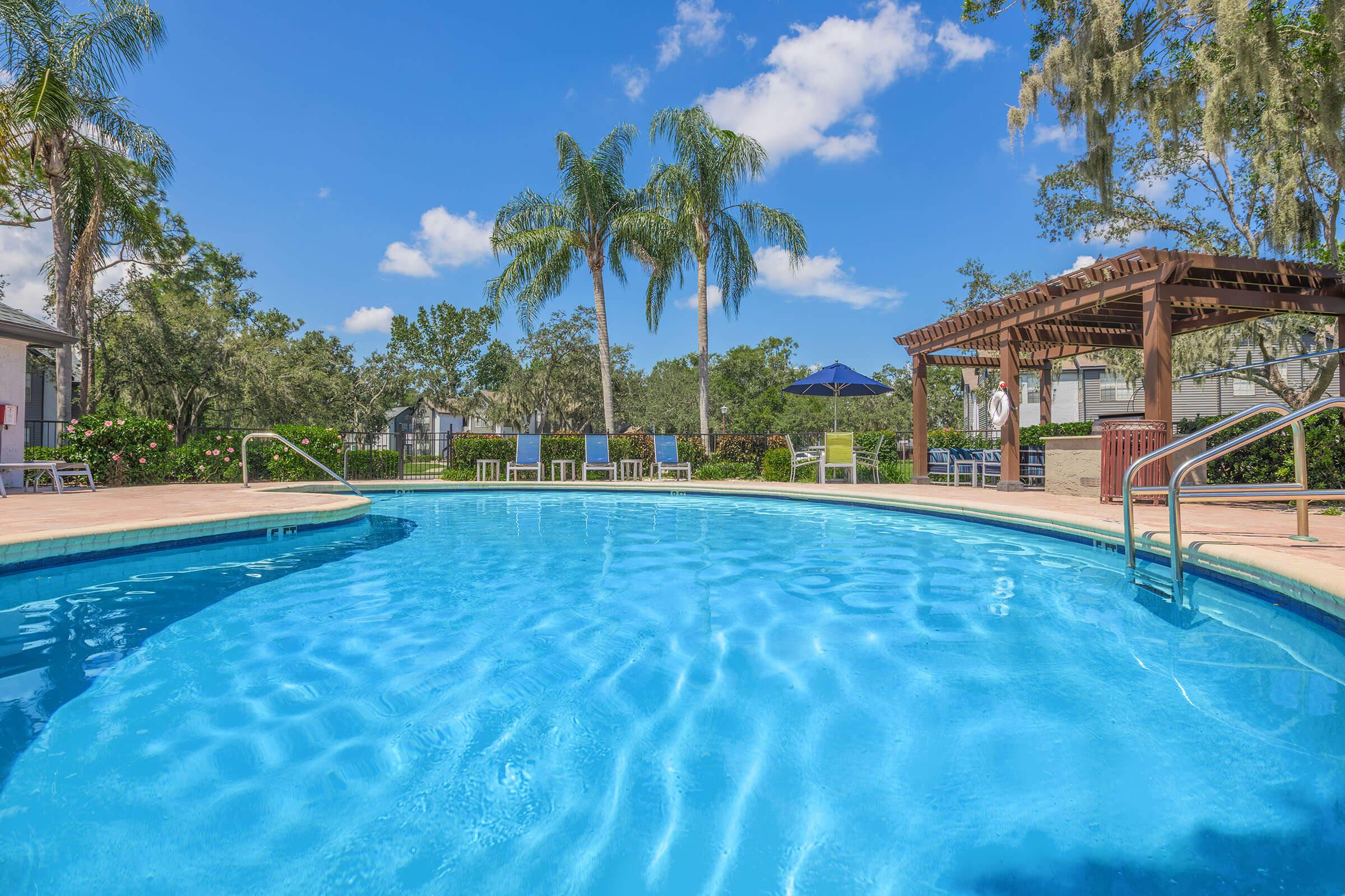 A clear blue swimming pool surrounded by lush greenery and palm trees, with a wooden pergola and lounge chairs nearby. The sky is bright with few clouds, creating a sunny, inviting atmosphere perfect for relaxation and leisure.