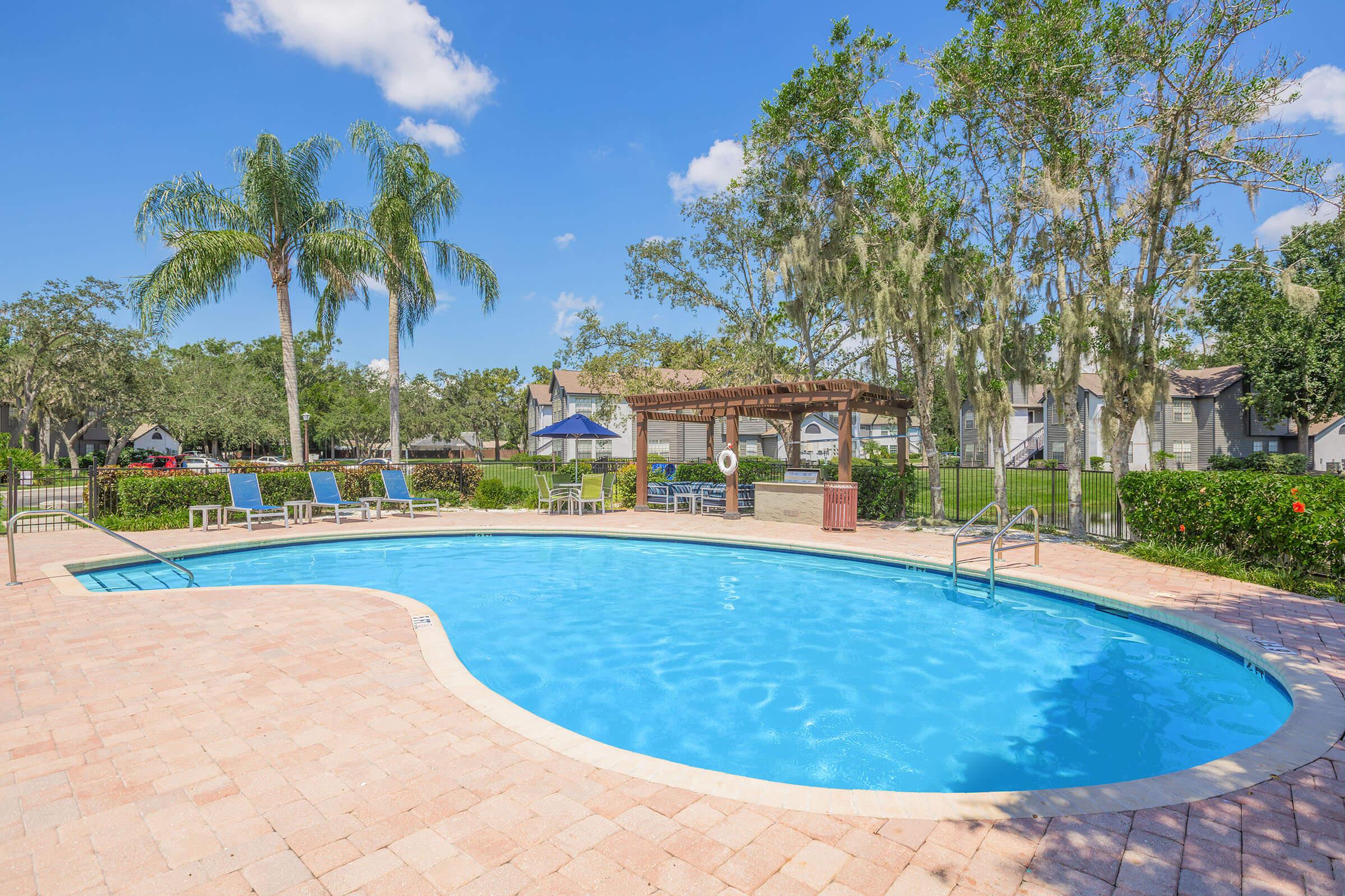 A clear blue swimming pool surrounded by palm trees and lounge chairs on a sunny day. In the background, there are shaded seating areas and green grass, creating a relaxing outdoor atmosphere. The sky is bright with a few clouds, complementing the tranquil scene.