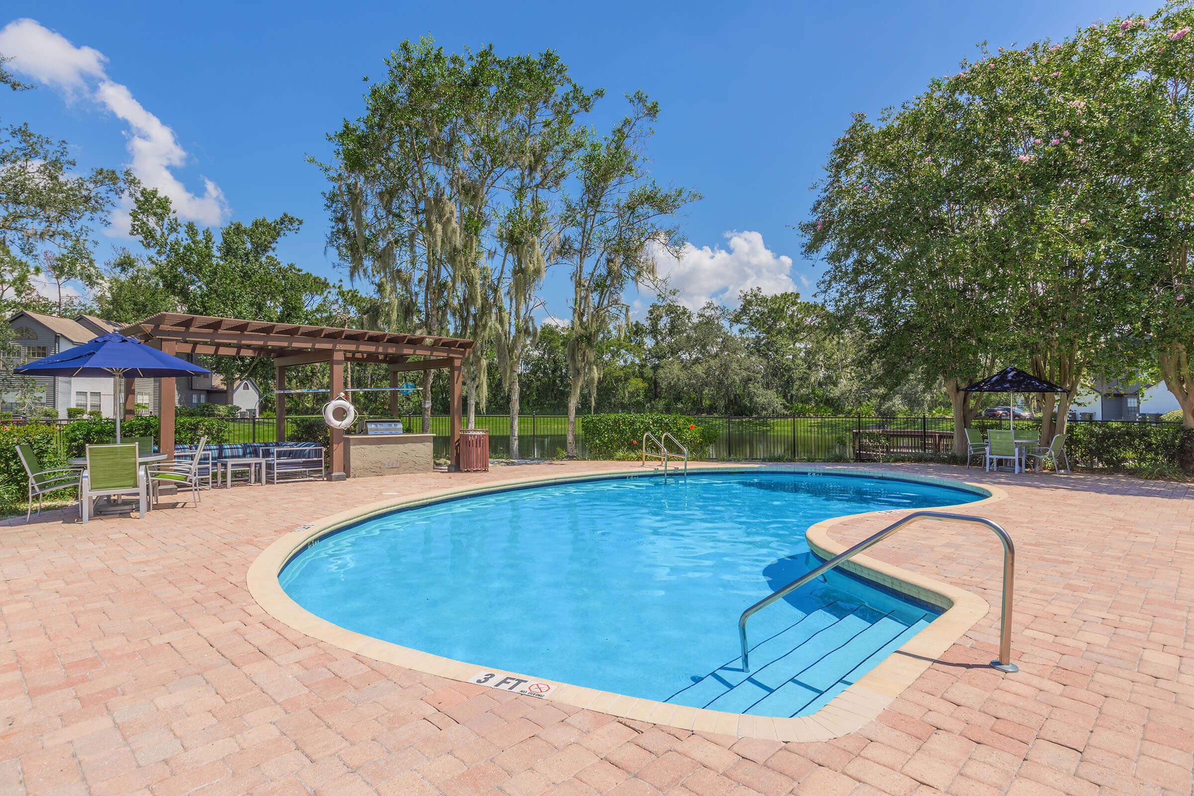 A bright outdoor swimming pool surrounded by lush greenery and trees. The pool features a curvy shape, a shallow area marked with "3 FT," and a metal ladder. There are lounge chairs and shaded seating areas nearby, with a pergola and blue sky filled with fluffy clouds in the background.