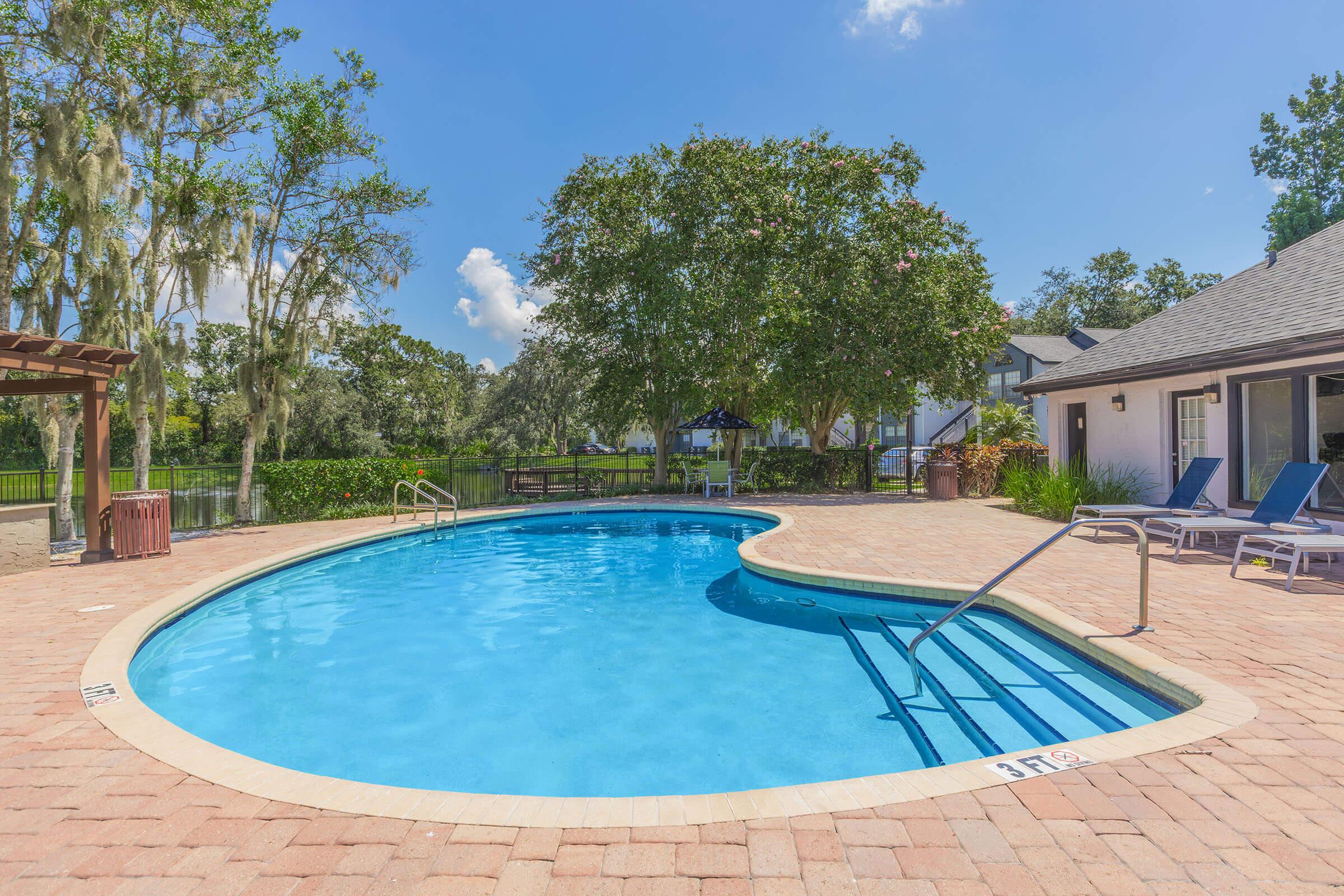A clear, inviting swimming pool with a curved edge, surrounded by a patio made of brick pavers. Sun loungers are placed nearby under a sunny sky with a few clouds. Lush greenery and trees frame the area, creating a relaxing atmosphere. A shaded seating area can be seen in the background.