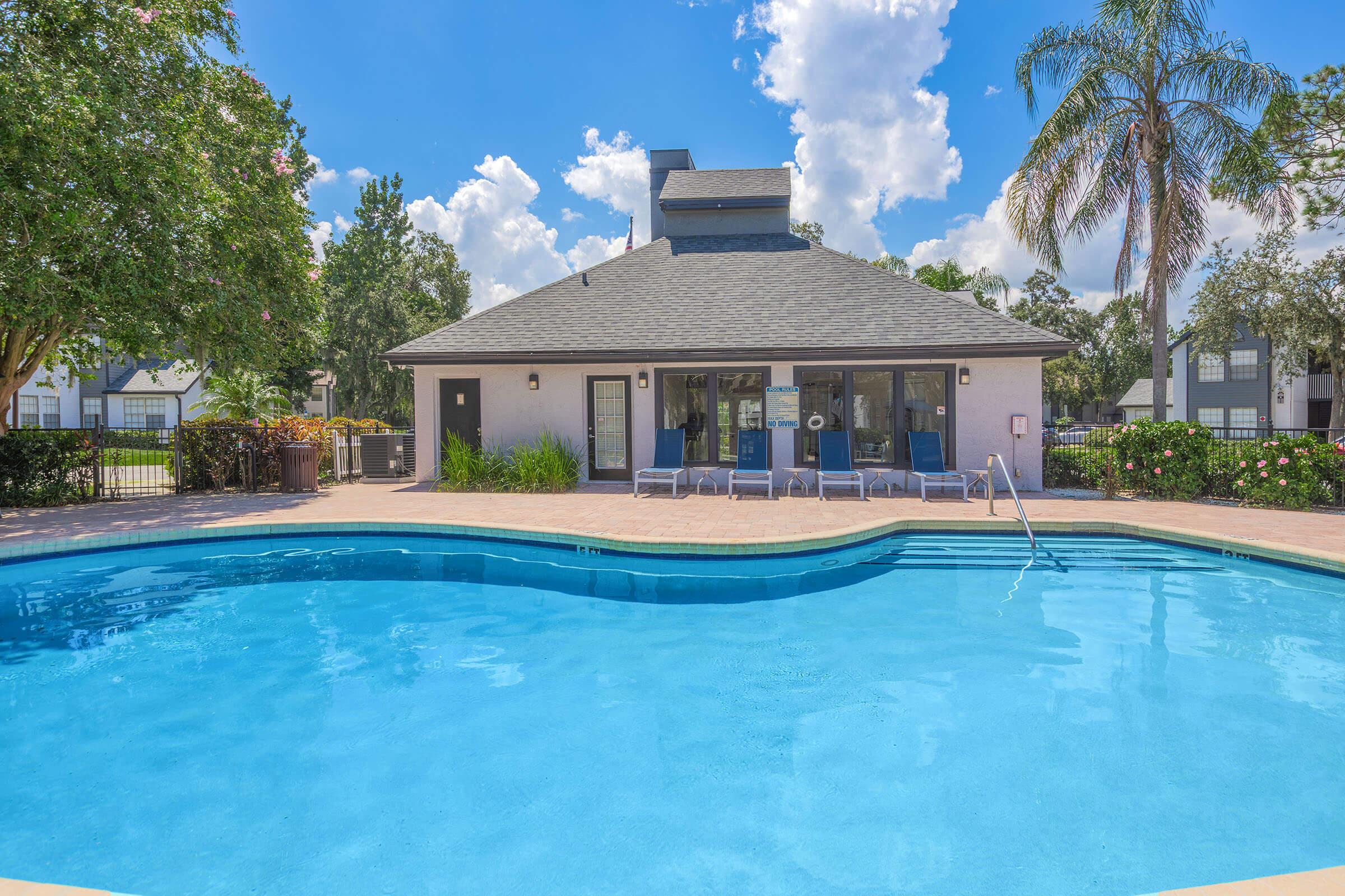 A clear blue swimming pool surrounded by lounge chairs, with a nearby clubhouse featuring large windows. Lush greenery and palm trees are visible, set against a bright blue sky filled with fluffy clouds. The scene conveys a relaxing outdoor atmosphere ideal for leisure.