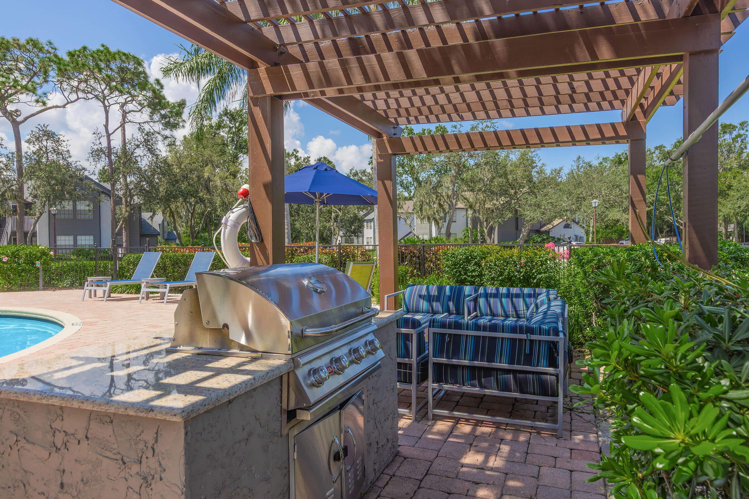 Outdoor grilling area near a swimming pool, featuring a stainless steel barbecue grill under a wooden pergola. Nearby, there are blue-striped seating arrangements, lounge chairs, and an umbrella, surrounded by lush greenery and a peaceful residential backdrop.
