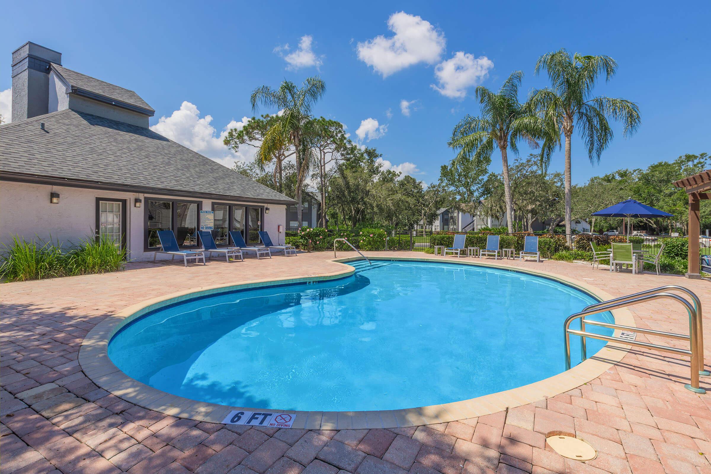 A sparkling circular swimming pool surrounded by paver stones, with lounge chairs nearby. In the background, a modern building with large windows and palm trees. The sky is bright blue with a few fluffy clouds, creating a vibrant and inviting atmosphere.