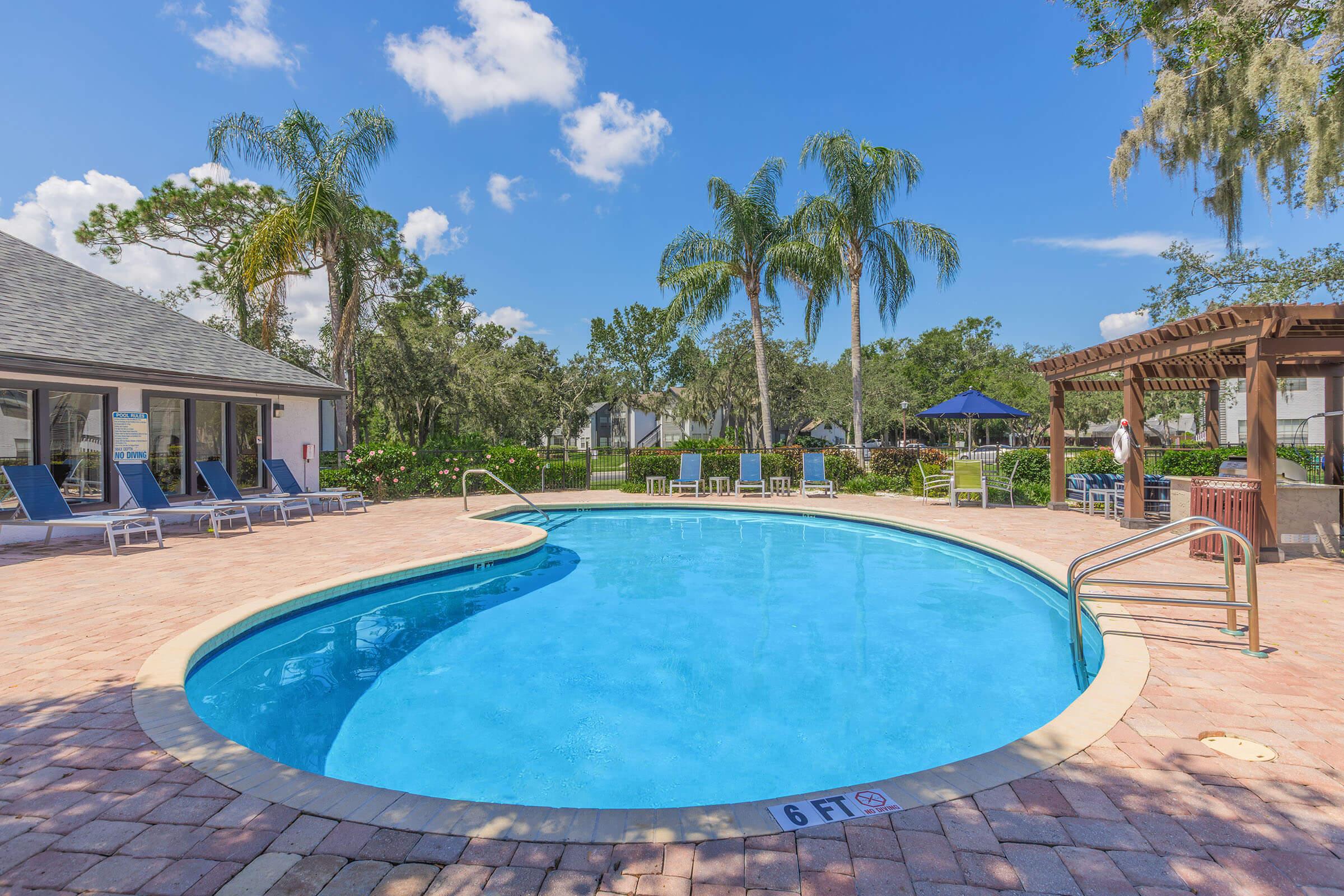 A clear swimming pool on a sunny day, surrounded by palm trees and lounge chairs. Nearby, there is a shaded area with a gazebo and additional seating. The sky is bright blue with a few clouds, creating a tranquil and inviting atmosphere.