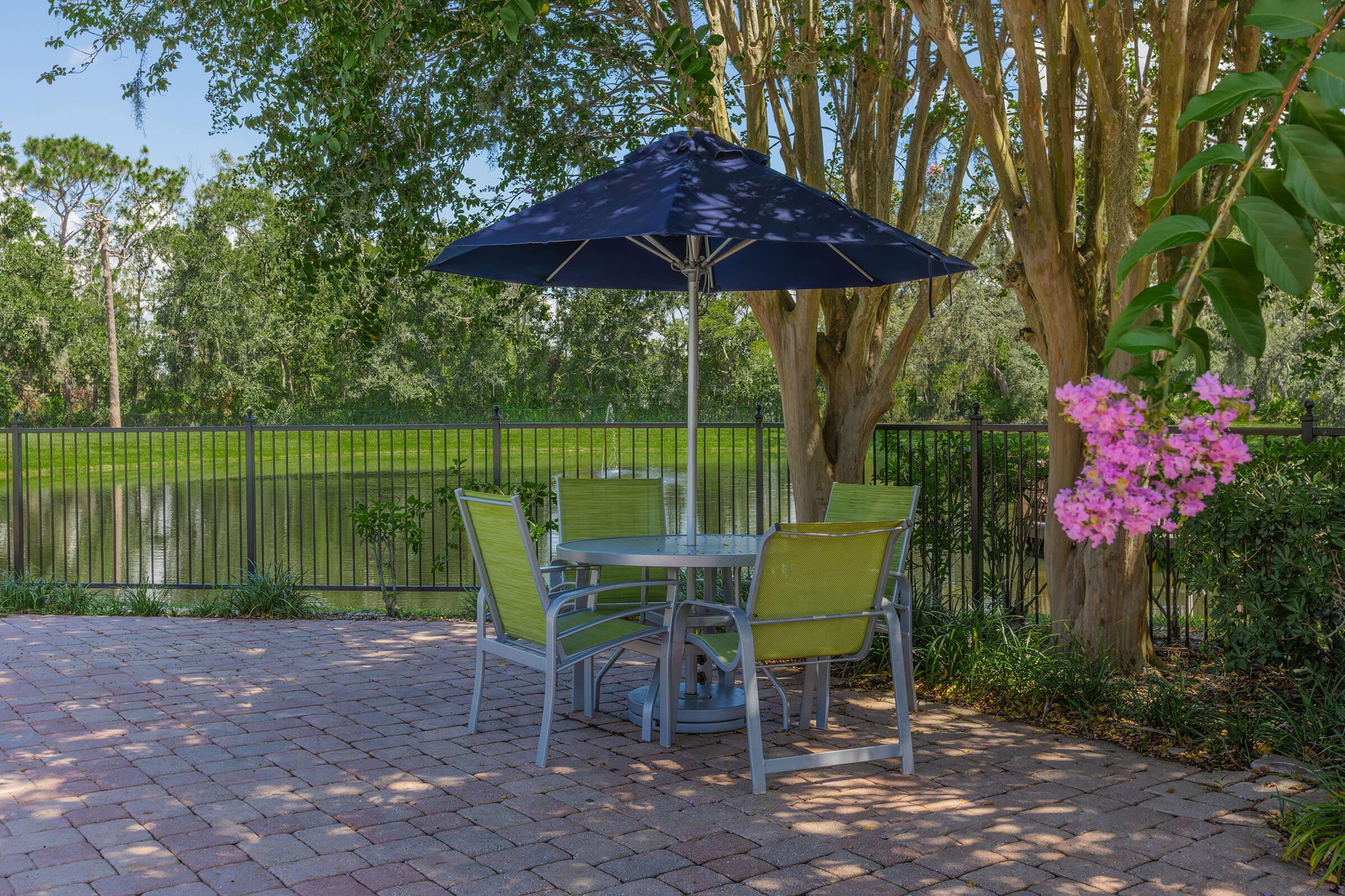 A patio area featuring a round table with four green chairs under a large black umbrella. Surrounding the space are lush trees and a flowering plant, and behind the patio is a serene lake bordered by a metal fence. The setting is bright and inviting, perfect for outdoor relaxation.