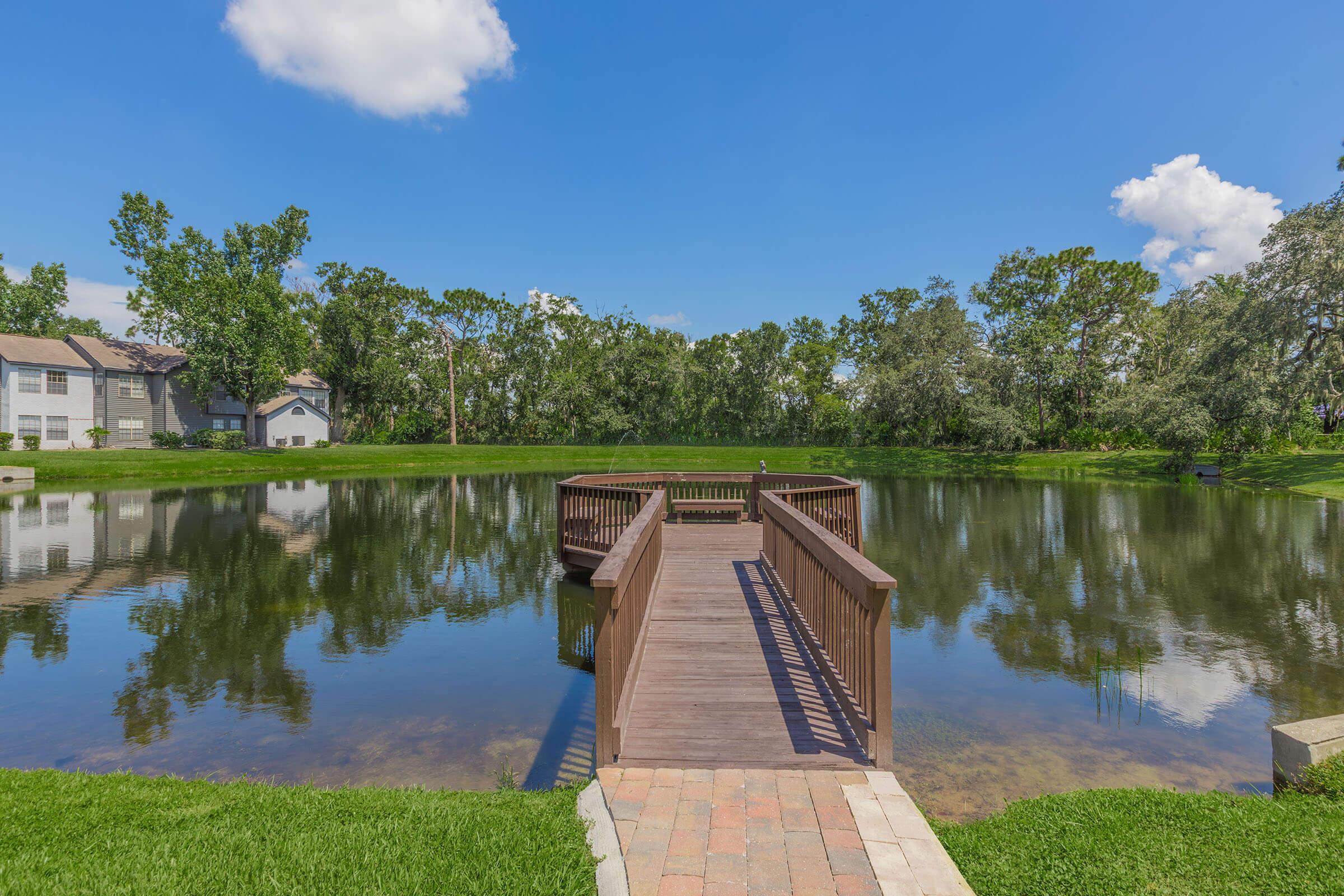 A peaceful scene featuring a wooden dock extending into a calm pond surrounded by lush greenery. The clear blue sky is dotted with a few clouds, and residential buildings are visible in the background, adding to the tranquil atmosphere of the setting.