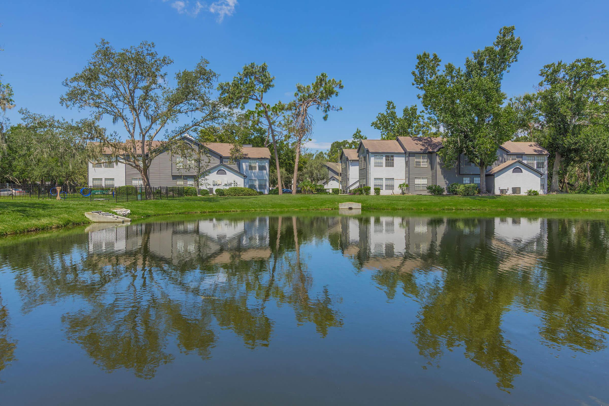 A peaceful residential area with modern buildings reflected in a calm pond. Lush greenery surrounds the water, with a clear blue sky overhead. The scene conveys tranquility and nature, ideal for leisurely strolls or relaxing by the water.