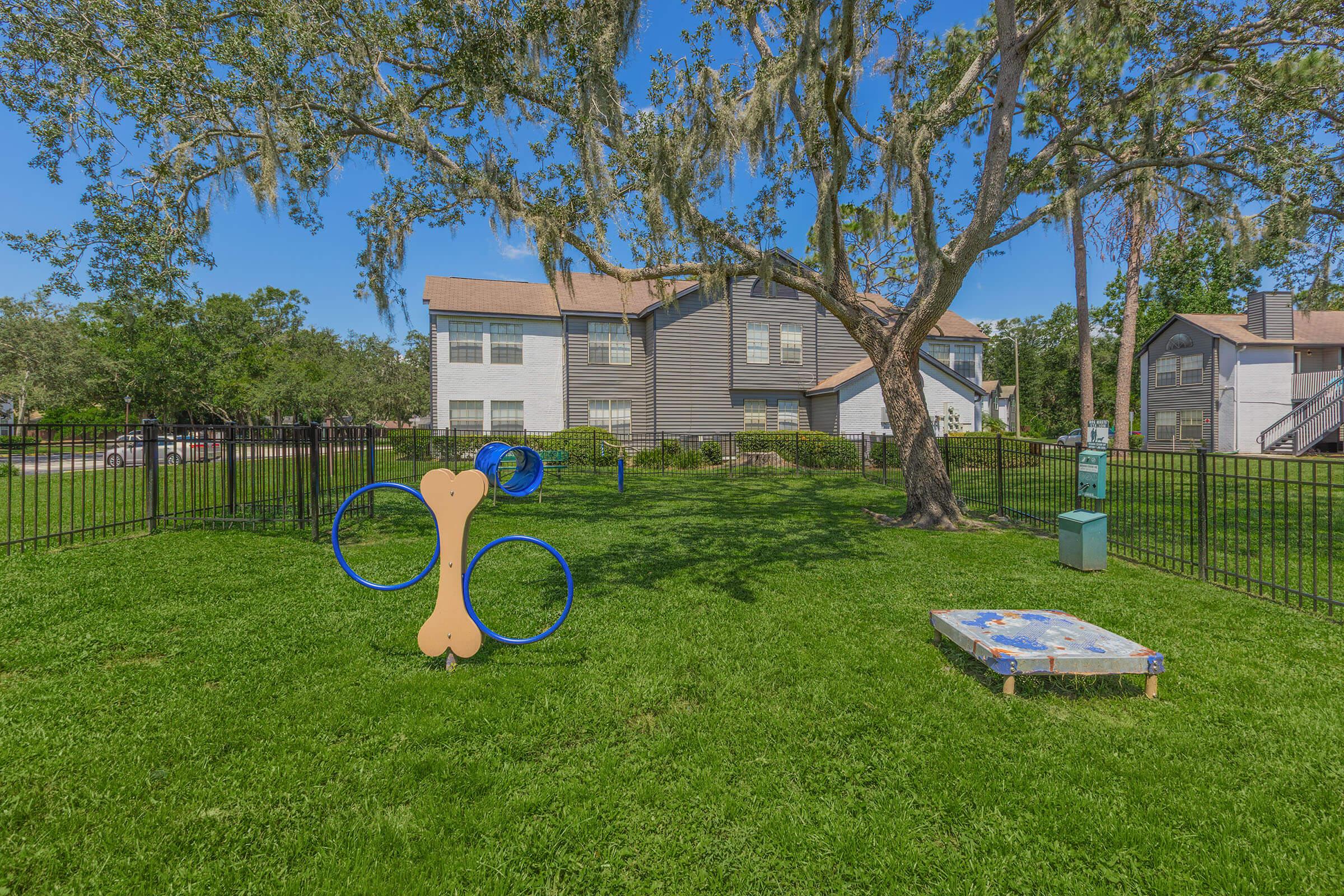 A grassy play area featuring a large tree and a fenced yard. There are two playful structures: a blue hoop set and a large dog bone-shaped frame. A small pet-friendly space with a raised platform is visible, situated near residential buildings in the background.