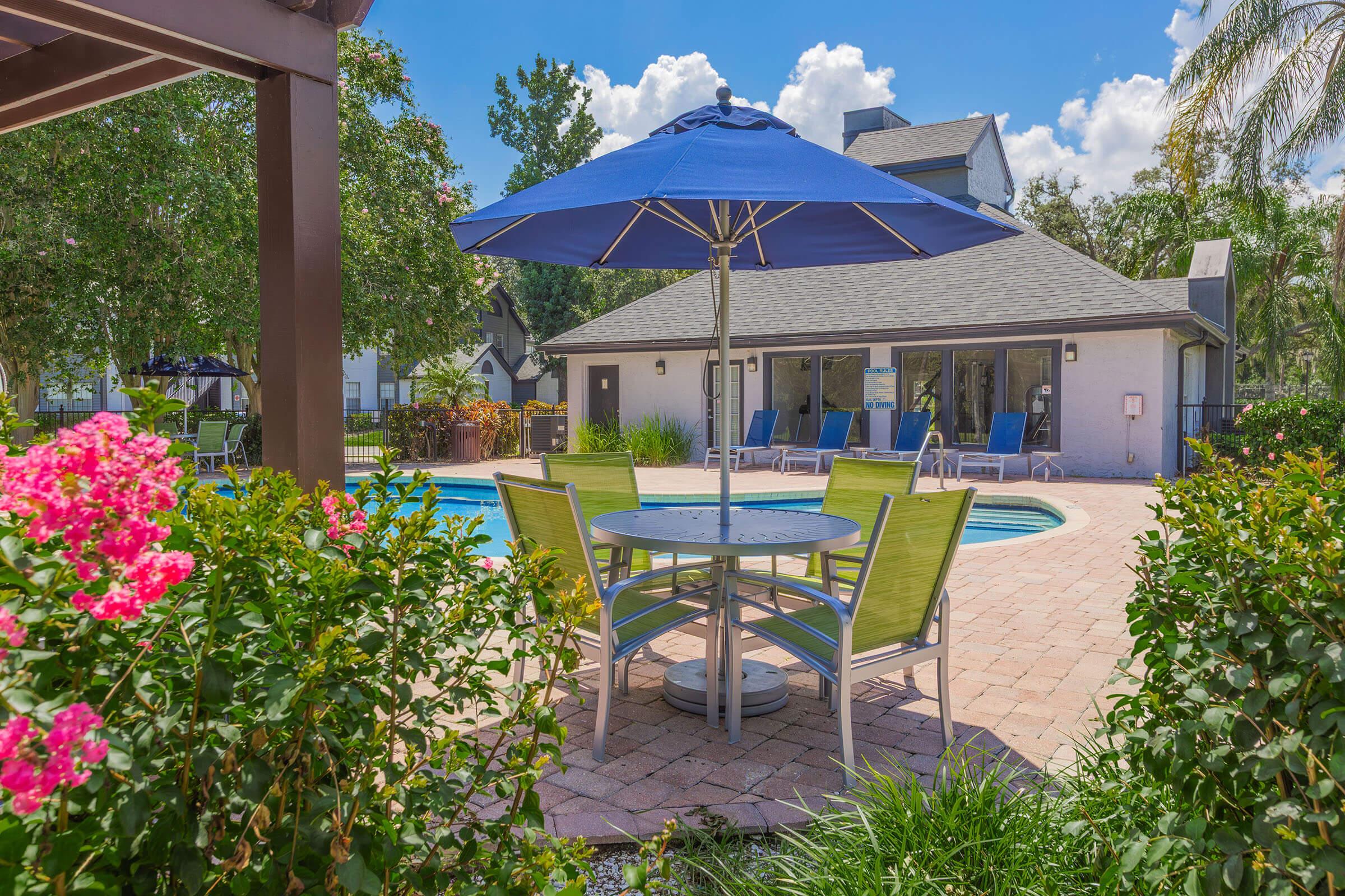 A sunny outdoor area featuring a sparkling swimming pool surrounded by green grass and vibrant flowers. A circular table with four brightly colored chairs is positioned under a large blue umbrella, with a clubhouse visible in the background and lounge chairs lining the poolside.