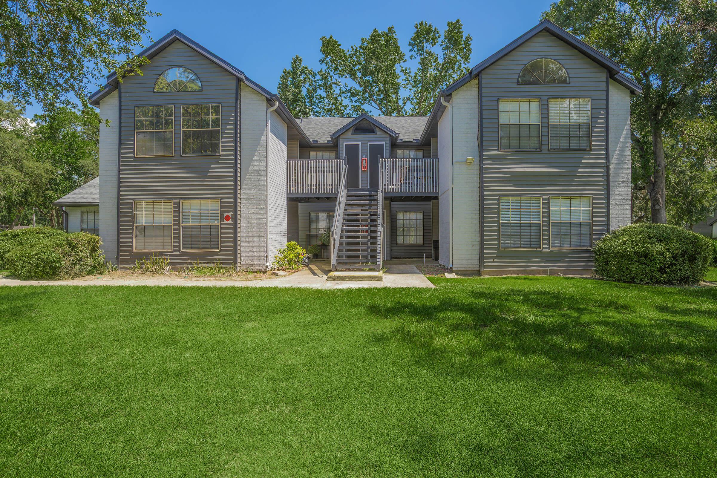 Two-story apartment building with a grey and white exterior, featuring large windows and a central staircase. The surrounding area is landscaped with green grass and shrubs, and there are trees in the background under a clear blue sky.