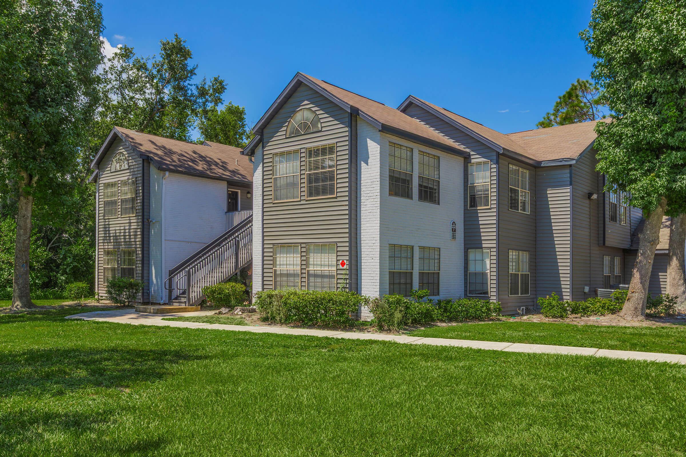 A well-maintained two-story apartment building with a grey exterior and large windows, surrounded by green grass and trees. The building features a small walkway leading to an entrance and a stairway on one side, under a clear blue sky.
