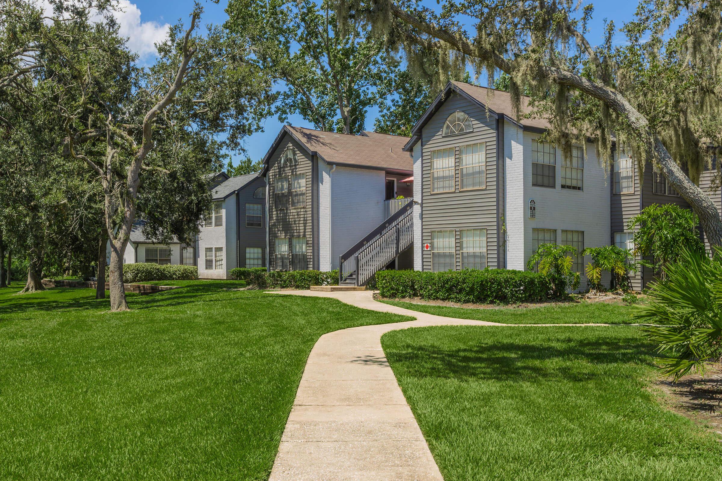 A well-maintained apartment complex with two-story buildings surrounded by lush green lawns and trees. A smooth concrete walkway leads through the landscaping, providing access to the apartments. The sky is clear with a few clouds, creating a pleasant and inviting atmosphere.