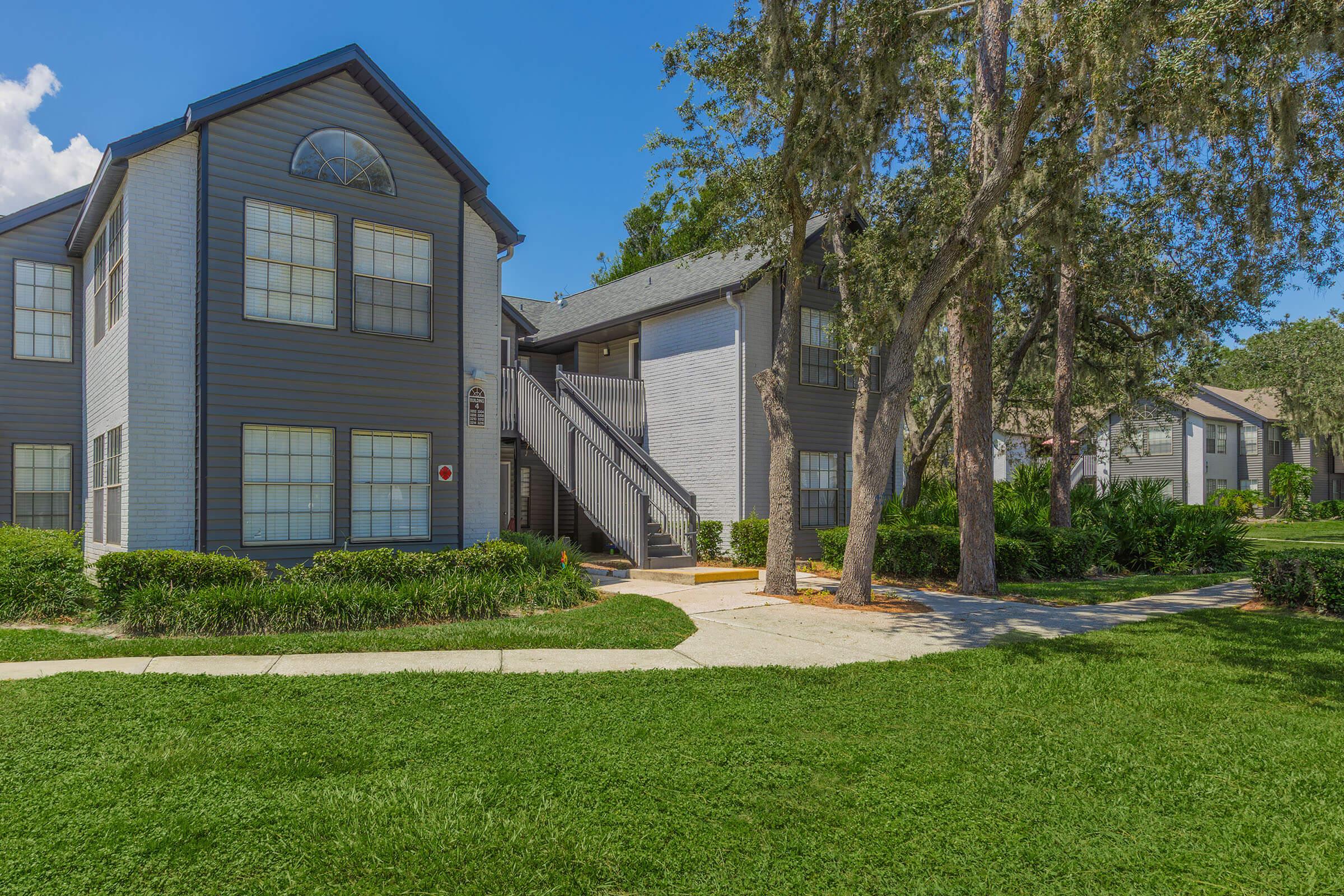 Two-story residential building with large windows and a gray exterior. The building features stairs leading to the second floor and is surrounded by green grass and shrubs. Tall trees are present nearby, under a clear blue sky. A well-maintained pathway leads to the entrance.