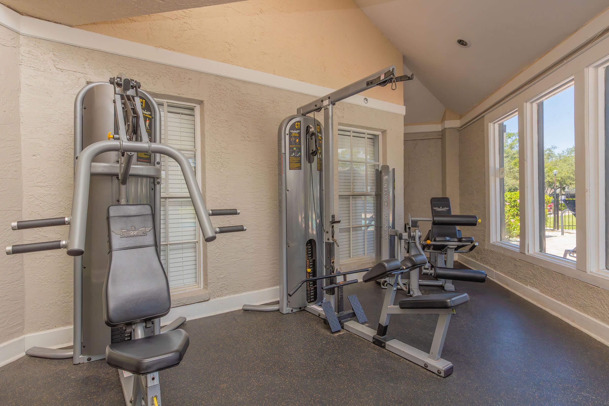 A well-lit gym area featuring various exercise equipment, including a multi-station weight machine and a bench press. Large windows allow natural light to fill the space, enhancing the workout environment. The floor is covered with dark, durable flooring suitable for fitness activities.