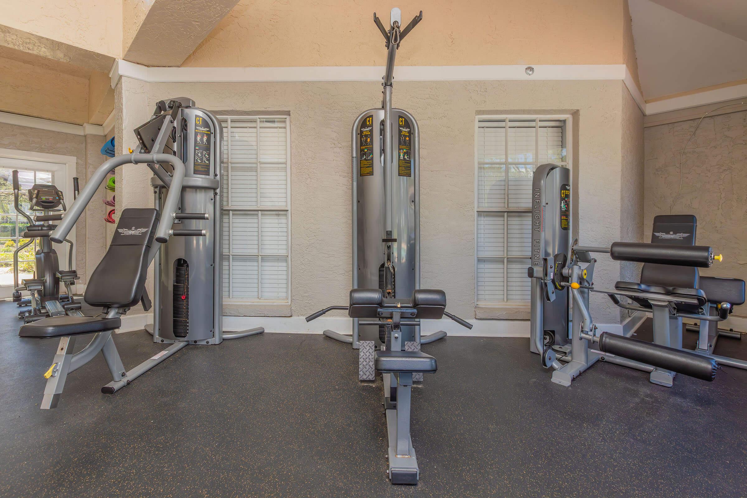 A well-equipped gym featuring various exercise machines, including a multi-station weight machine, a bench press, and a cardio machine. The space is bright with large windows illuminated by natural light, and the flooring is black rubber for safety and comfort.