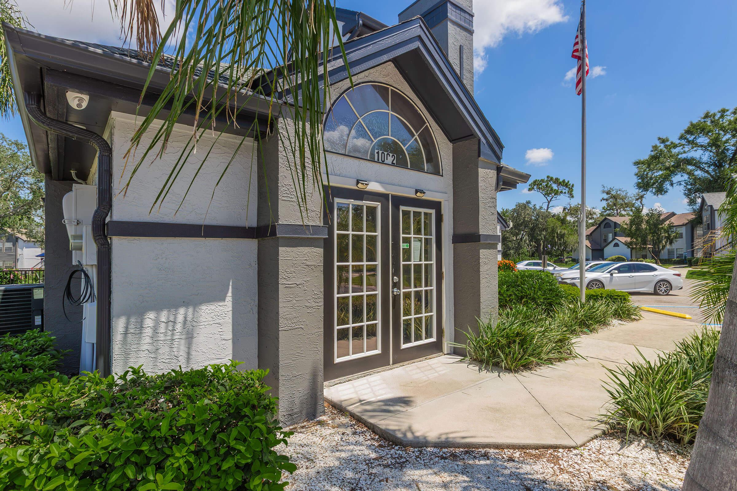 Exterior view of a modern building featuring large double doors with a rounded top, surrounded by greenery. A flagpole with an American flag is visible, along with a parking area in the background. The scene is bright and sunny, showcasing a well-maintained entrance.