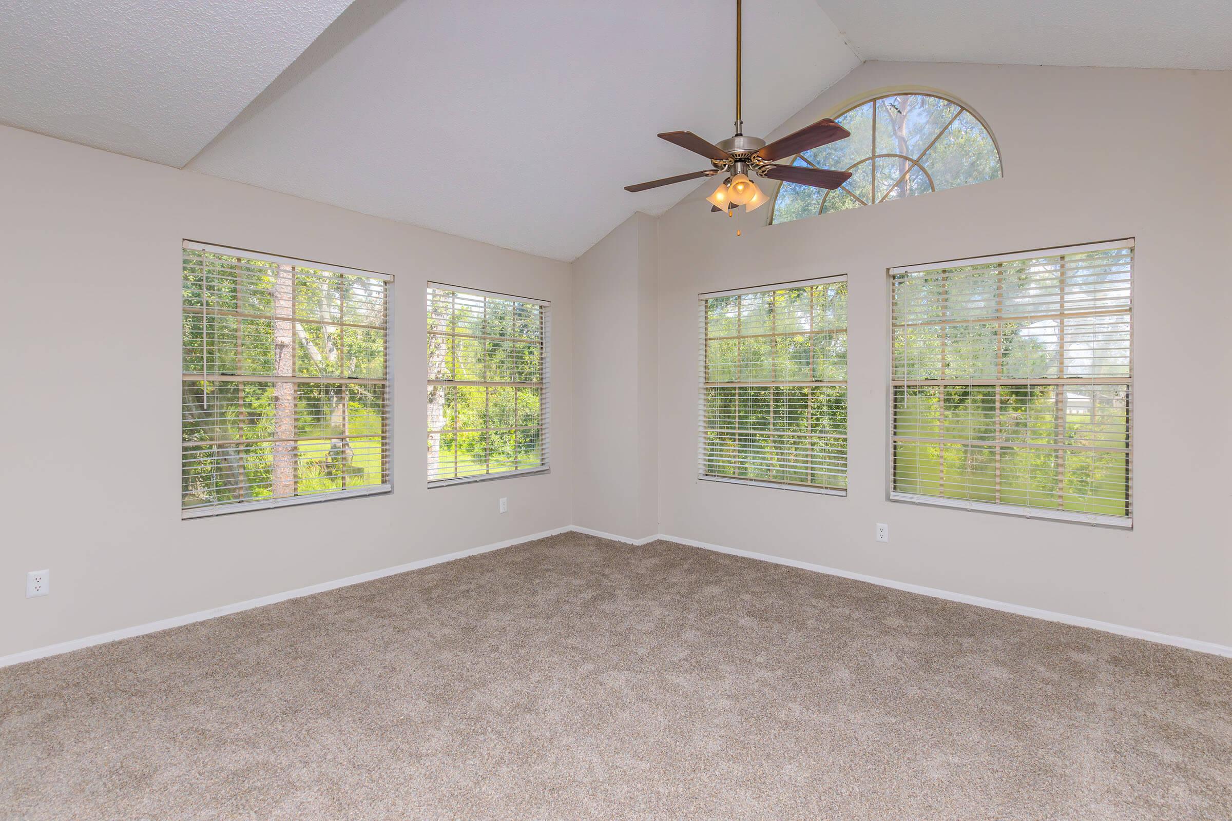 A spacious room with a high ceiling and large windows featuring blinds. The walls are painted a light color and the floor is covered in tan carpet. A ceiling fan is mounted in the center, and the view outside shows greenery, creating a bright and airy atmosphere.