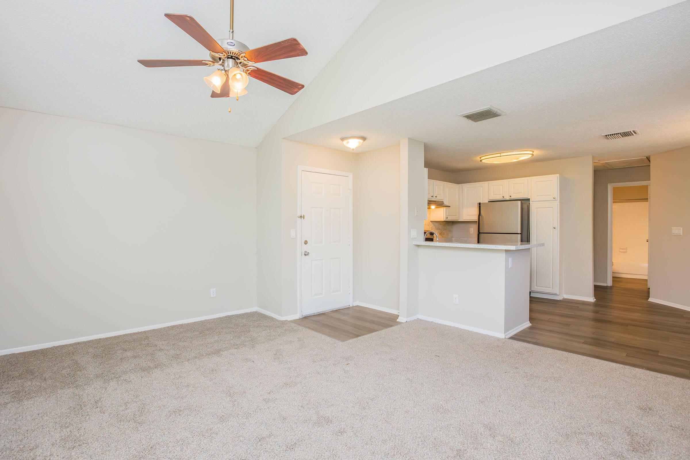 A spacious living area with a ceiling fan, light fixture, and beige carpet. The entrance leads to a kitchen with white cabinetry and stainless steel appliances in the background. Bright walls and a high ceiling create an open, airy atmosphere.