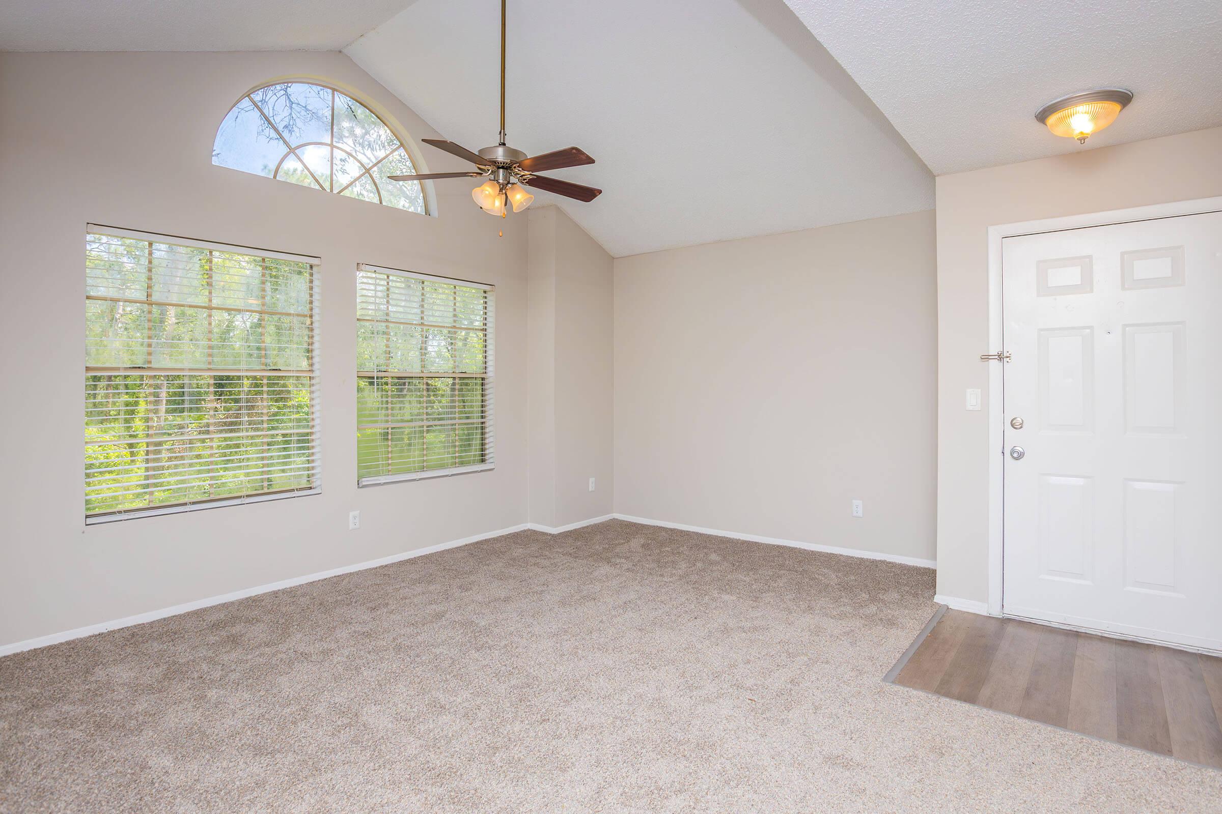 A spacious, empty living room featuring light beige carpet, a ceiling fan, and large windows with blinds allowing natural light. The room has a vaulted ceiling and a front door leading outside, with a small entryway area.