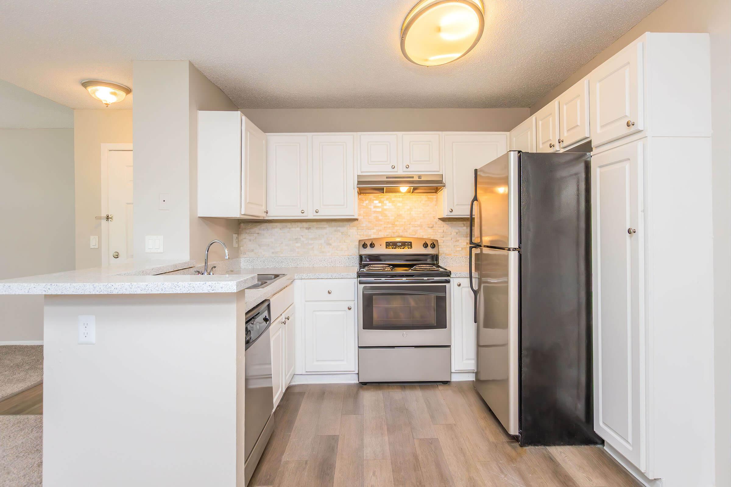 Modern kitchen featuring white cabinetry, stainless steel appliances, and a spacious layout. Includes a refrigerator, stove, and dishwasher, with a light-colored countertop and backsplash. The flooring is a light wood finish, creating a bright and inviting atmosphere.
