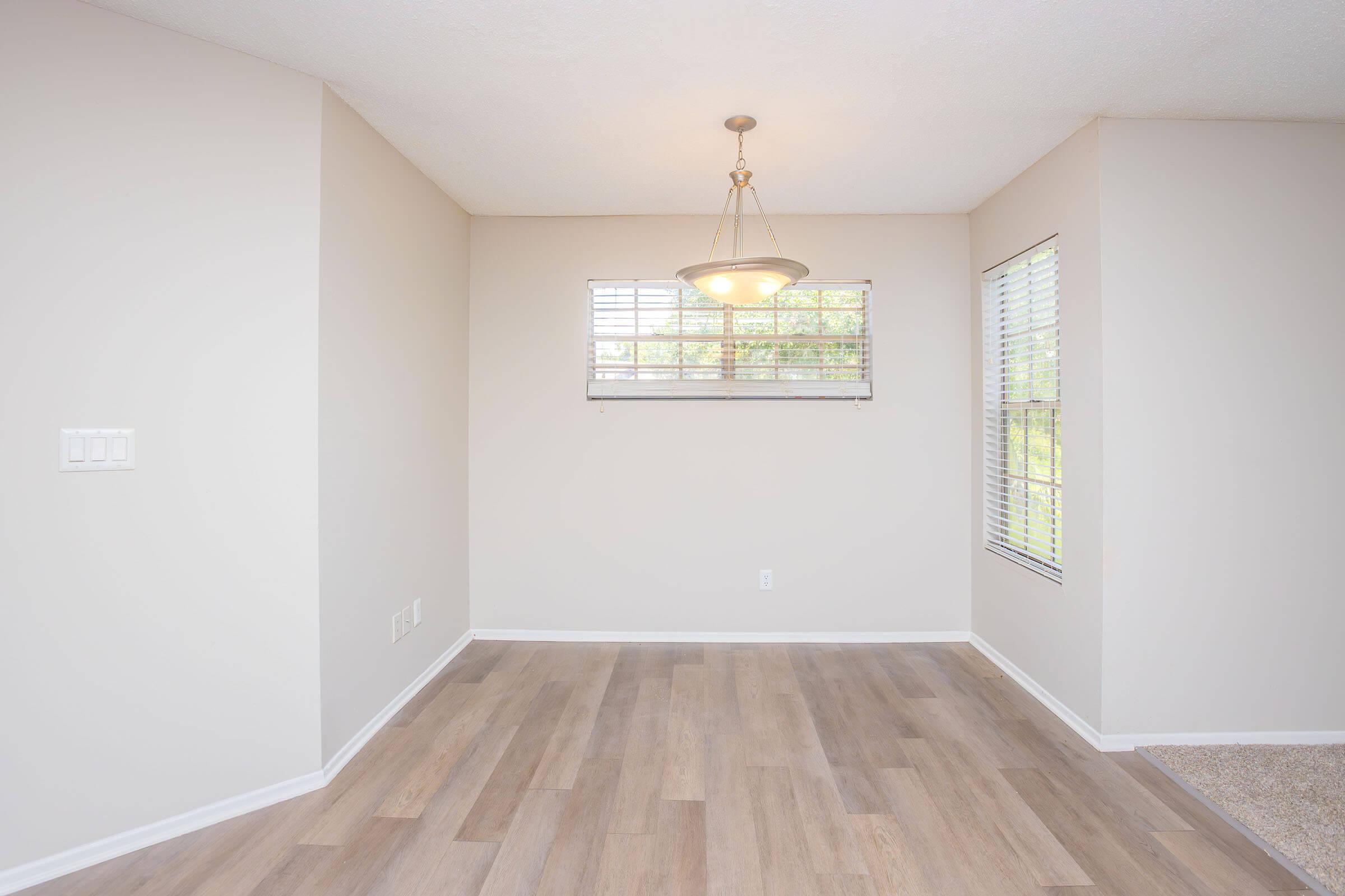A well-lit room with light-colored walls and a large window covered by blinds, letting in natural light. The floor features light wood laminate, and there's a small ceiling light fixture. One wall includes a panel with light switches. The space appears clean and empty, ready for furnishings.