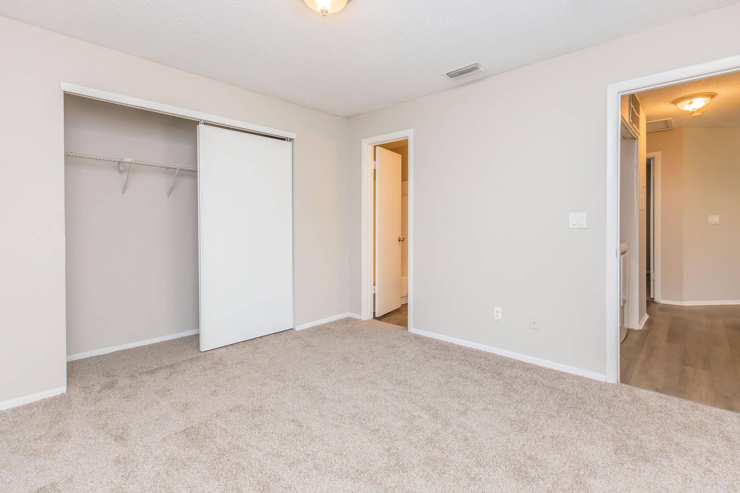 A spacious, empty bedroom with light-colored walls and carpeted flooring. It features a closet with sliding doors on the left, a doorway leading to another room on the right, and a ceiling light fixture. The room has a tidy and neutral aesthetic, ideal for personalization.