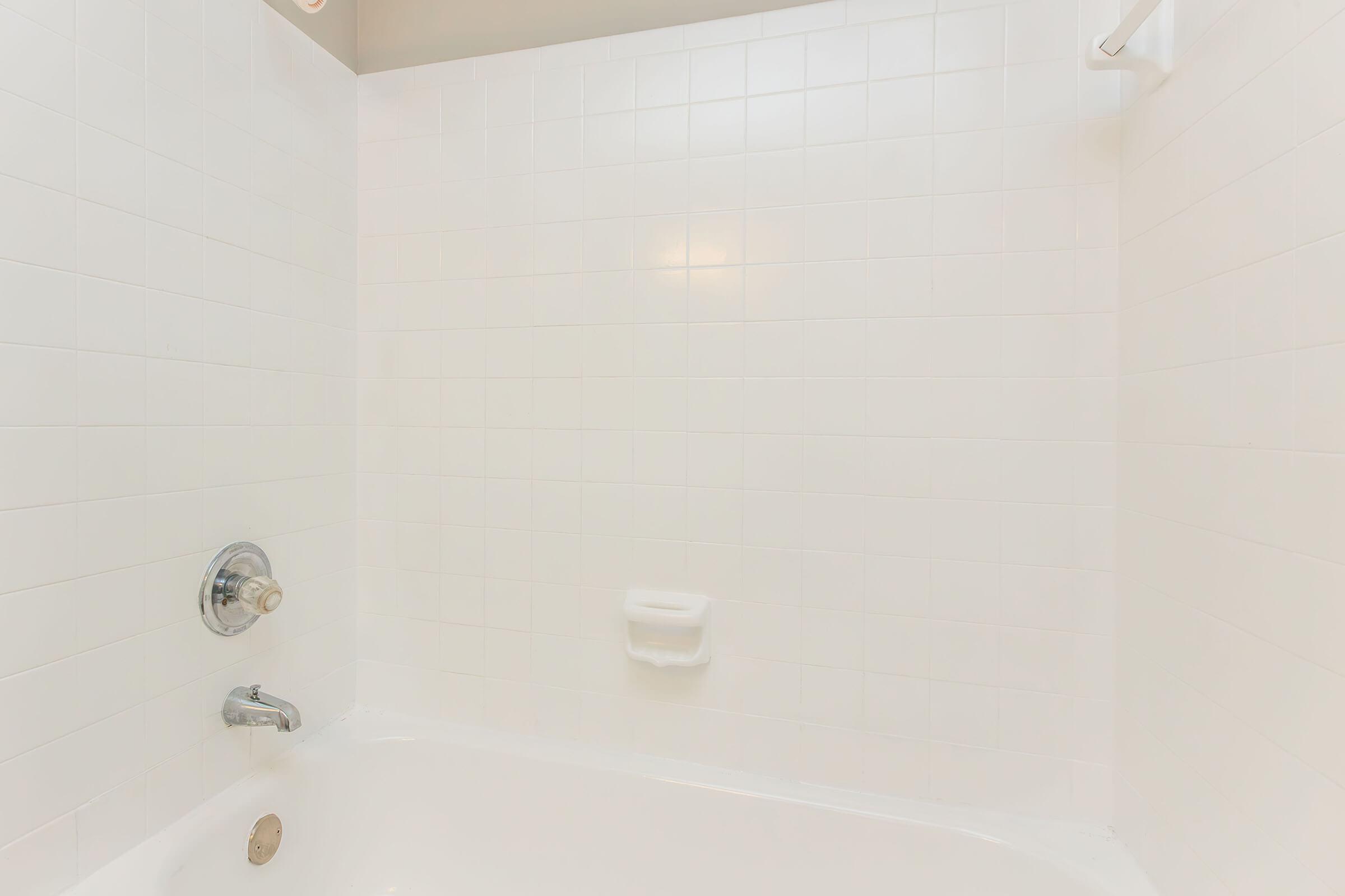 A clean, empty bathtub with white tiled walls. The tub features a chrome faucet and a handheld showerhead. There is a wall-mounted grab bar and a built-in soap dish. The overall aesthetic is bright and minimalistic, emphasizing a tidy and spacious bathroom environment.