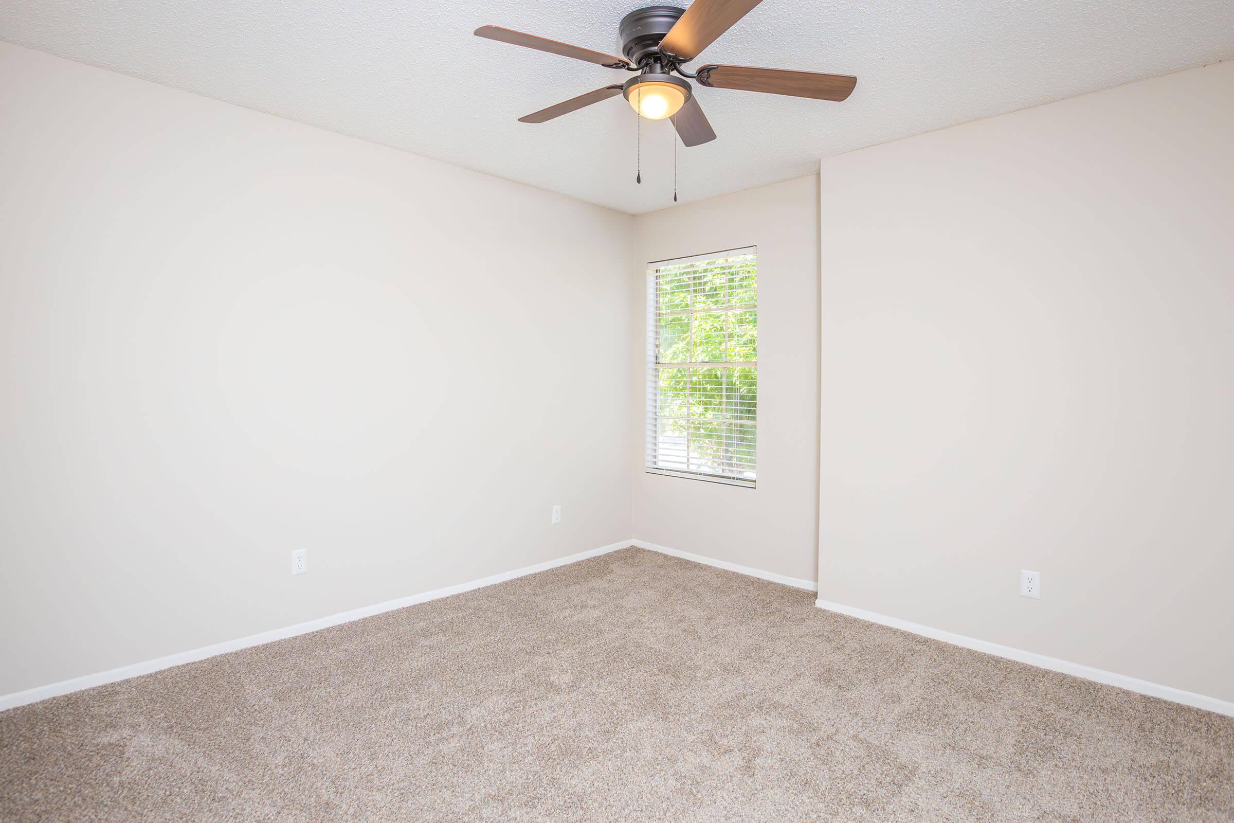 Empty room with light beige walls and a ceiling fan. The floor is covered in light brown carpet. There's a window with white blinds allowing natural light to enter. The space is well-lit and uncluttered, creating a bright and airy atmosphere.