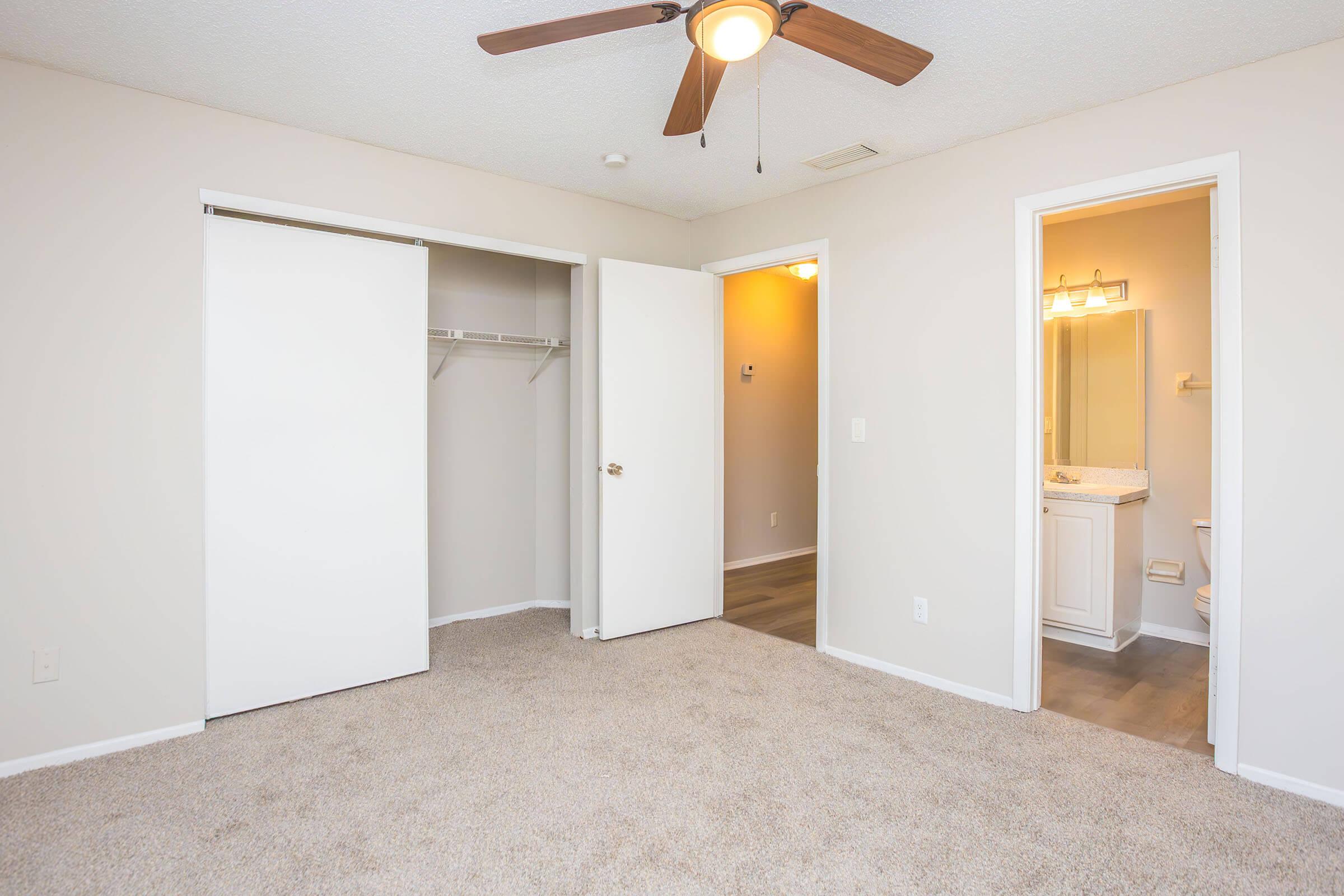 A spacious, light-colored bedroom featuring beige carpet, a ceiling fan, and double doors leading to a closet. To the right, there is an adjoining bathroom with a door partially open, revealing a vanity and bathroom fixtures. The walls are painted in a neutral shade, creating a calm atmosphere.