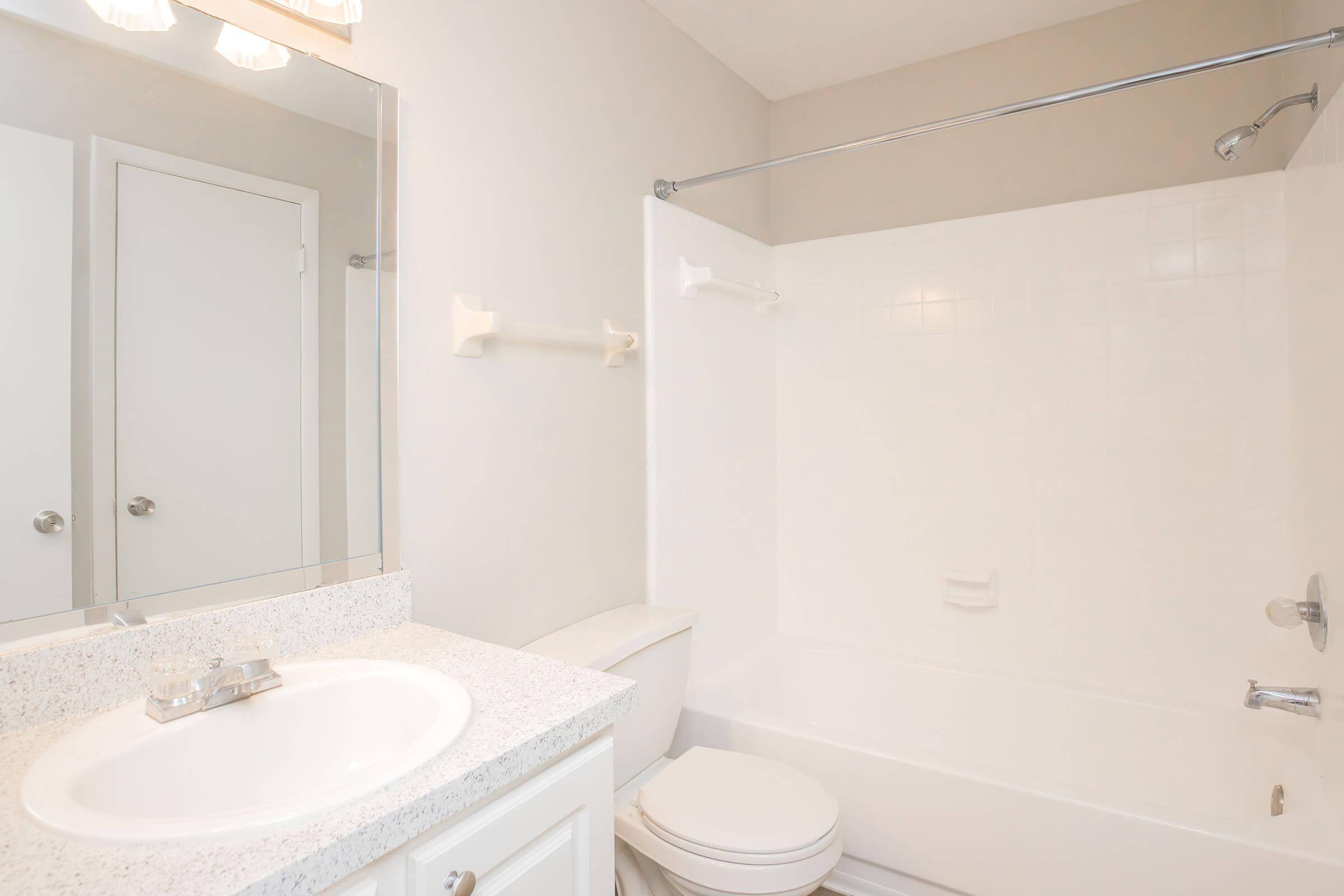 Bright, modern bathroom featuring a white bathtub and shower combination, a wall-mounted mirror above a granite countertop with a sink, a white toilet, and light-colored walls. The space is well-lit with overhead lighting and has a door leading to another room.