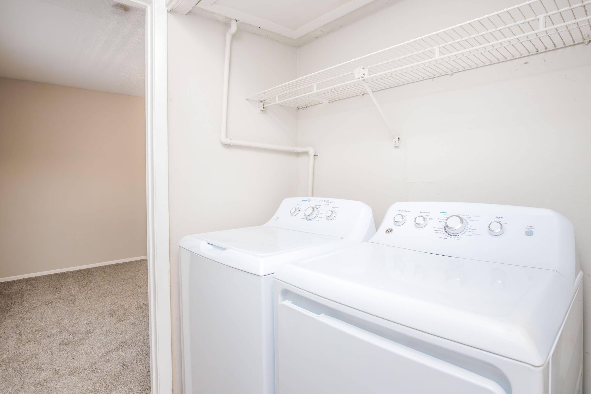 A laundry area featuring a white washing machine and a dryer placed side by side, with a wire shelf above them. The walls are light-colored, and the floor is carpeted in a neutral shade, creating a clean and organized look in the space.