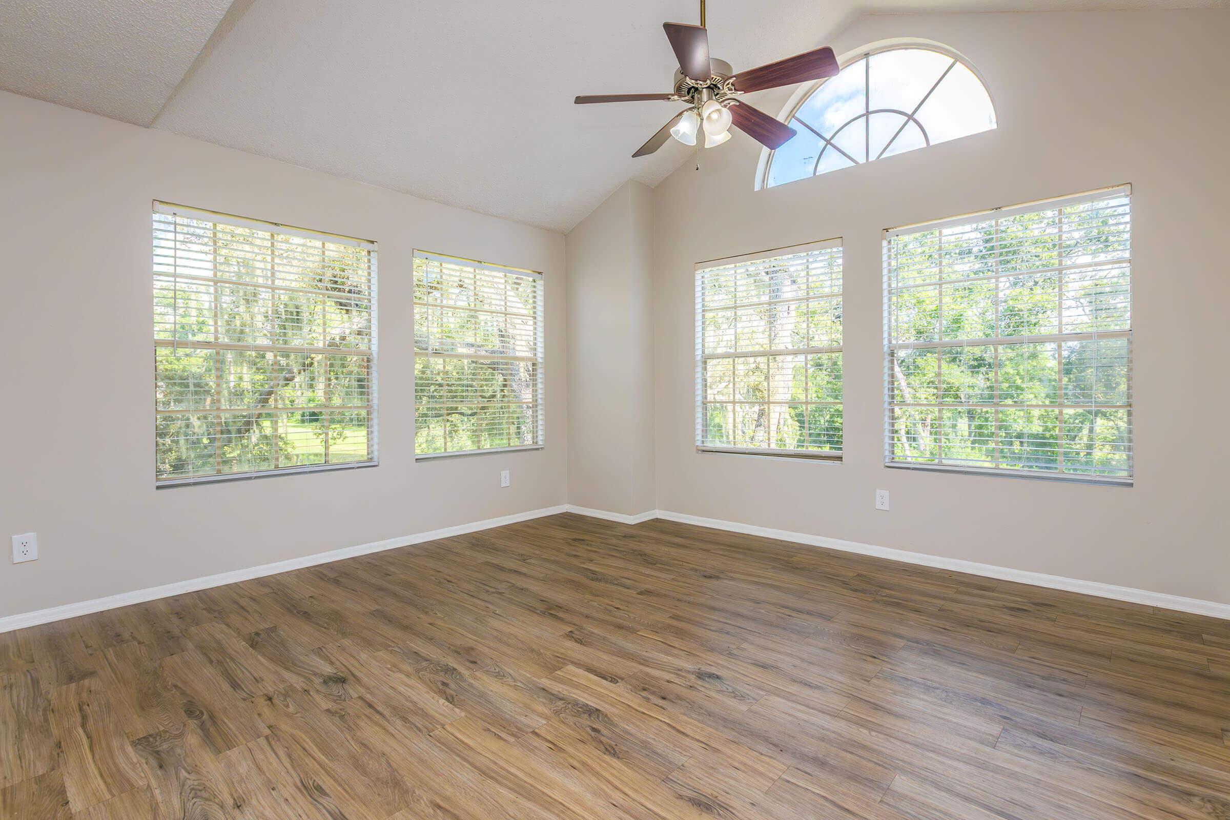 A spacious room with tall windows that allow natural light to fill the space. The room features a ceiling fan and a light wood floor, with neutral-colored walls and no furniture, creating a bright and open atmosphere. Greenery is noticeable outside the windows.