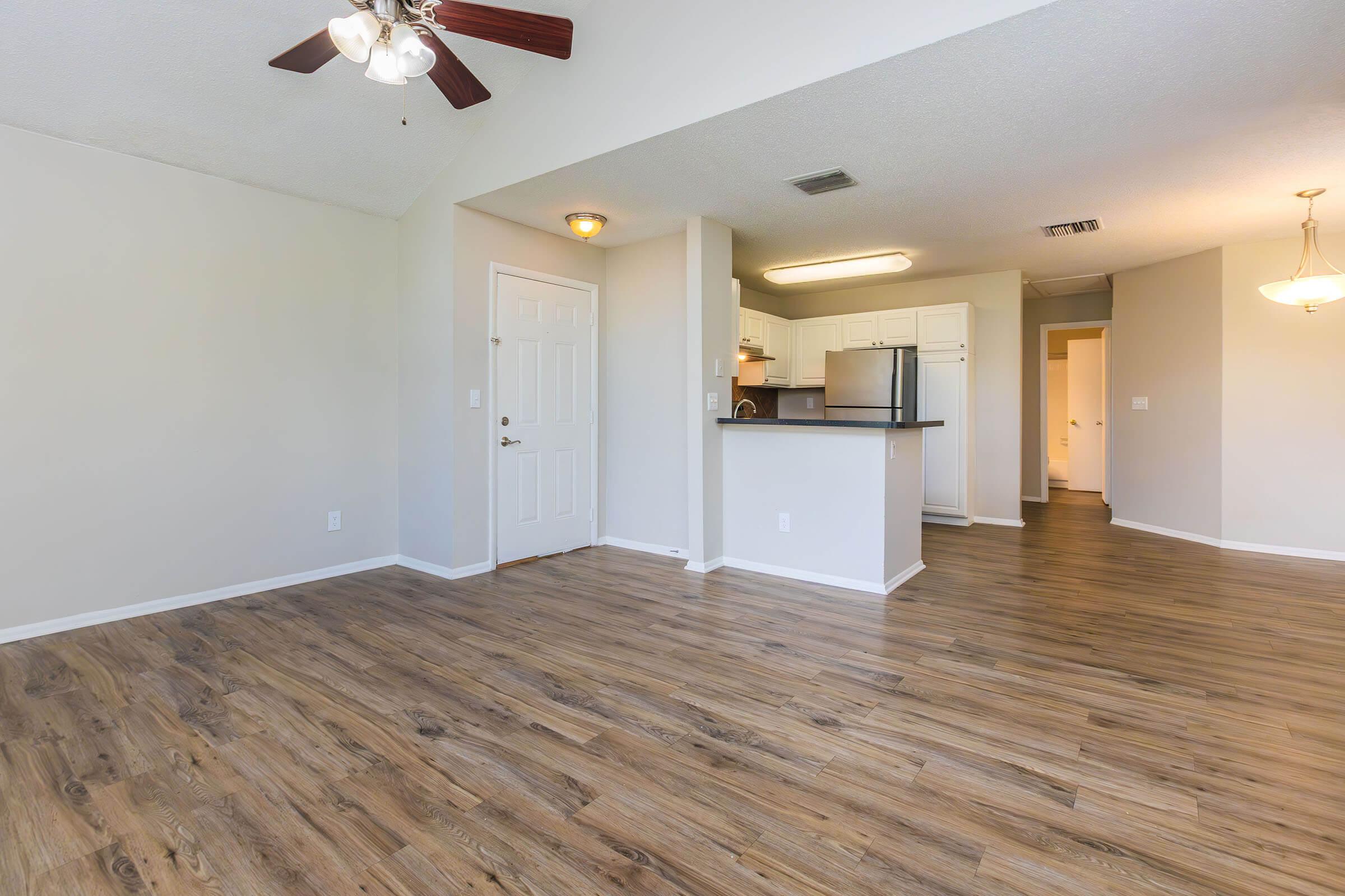 Interior view of a spacious, light-filled living area with hardwood-like flooring, a ceiling fan, and an open kitchen area in the background. The kitchen features white cabinets and appliances, with a doorway leading to an unseen area.