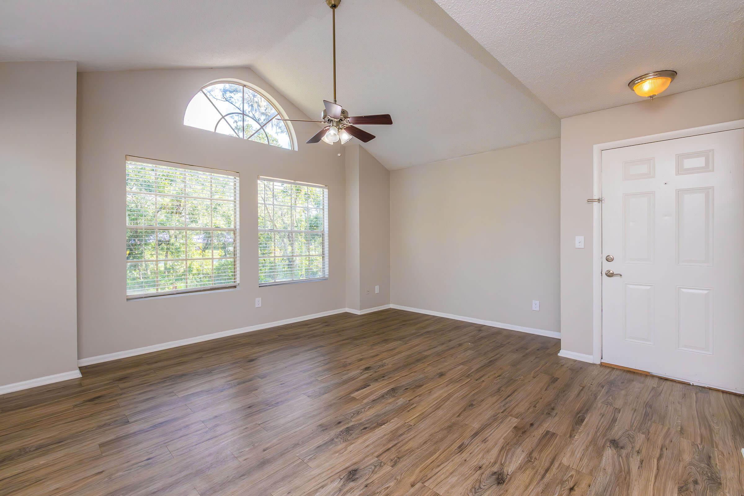 Spacious, well-lit living room featuring large windows with a decorative arch, ceiling fan, and hardwood-style flooring. The room is painted in neutral colors and includes a front door with a light fixture nearby.