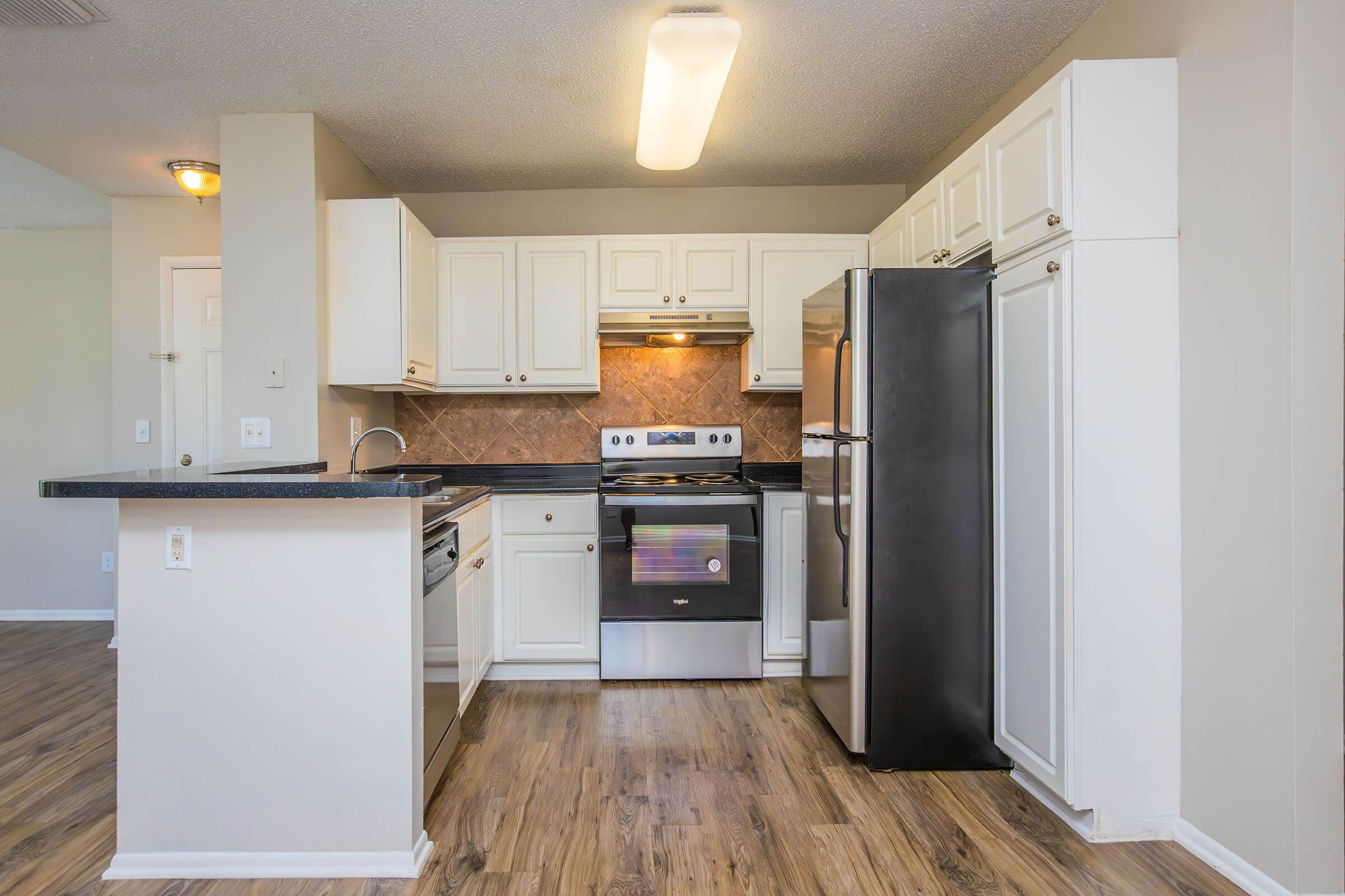 Modern kitchen featuring white cabinetry, stainless steel appliances including a refrigerator and stove, a black countertop, and wood-like flooring. The space is well-lit with natural and artificial light, showcasing a clean and functional design.