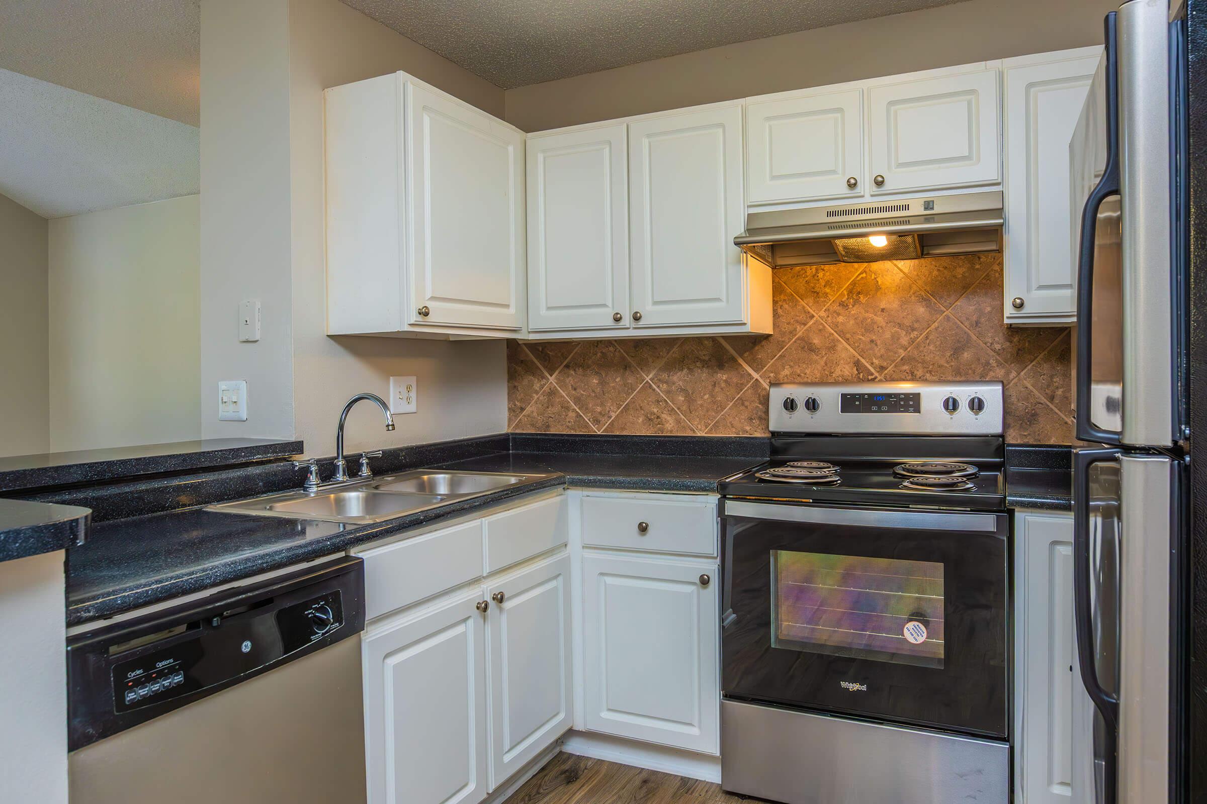 Modern kitchen featuring white cabinetry, a stainless steel refrigerator, black countertops, and a silver oven with a glass cooktop. The space includes a double sink and a dishwasher, with decorative tile backsplash and warm under-cabinet lighting.