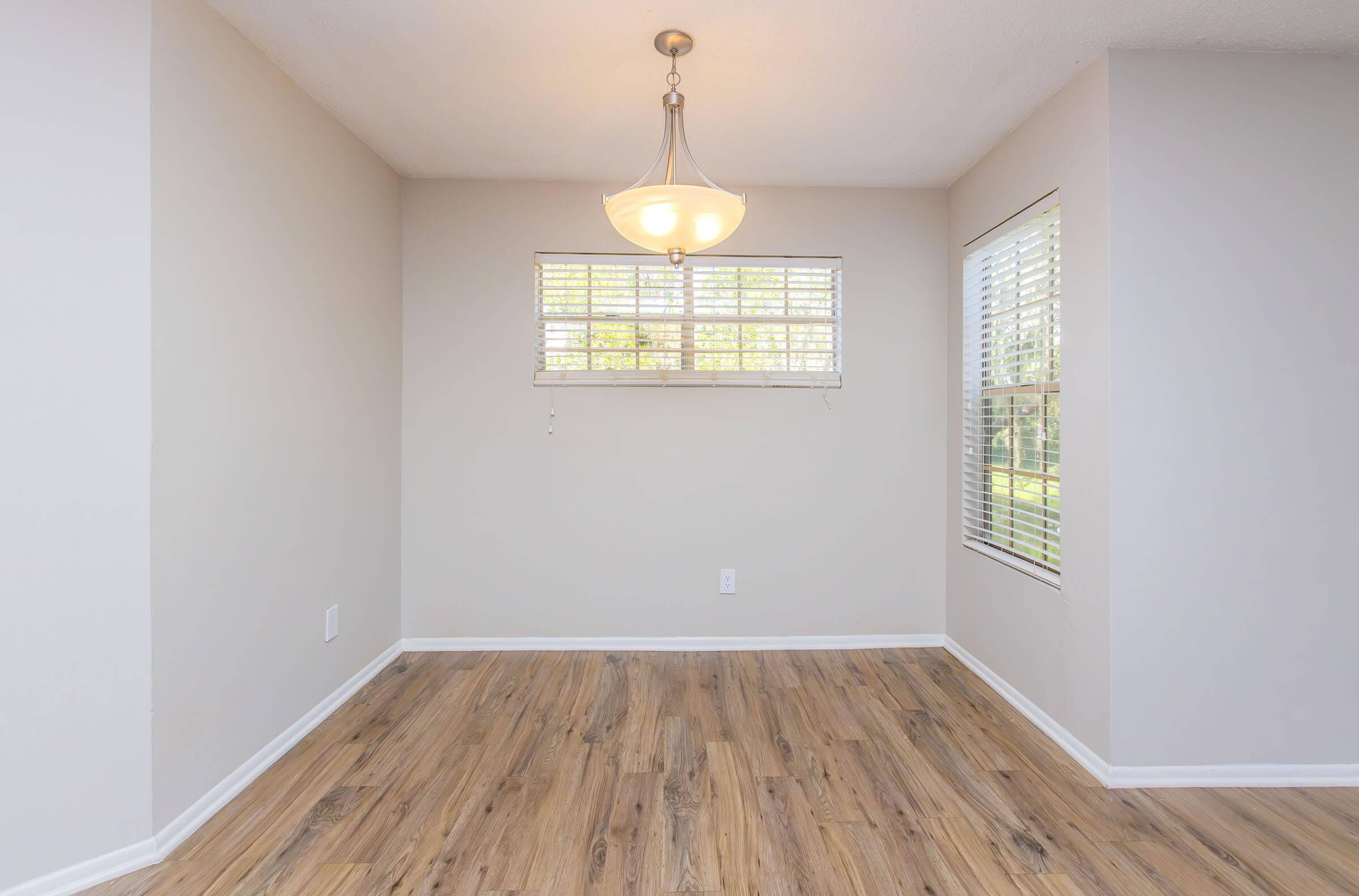A well-lit empty room with a light fixture hanging from the ceiling, featuring light-colored walls and wooden flooring. There are two windows with blinds, allowing natural light to enter, and a clean, spacious feel. Perfect for a dining or living area setup.