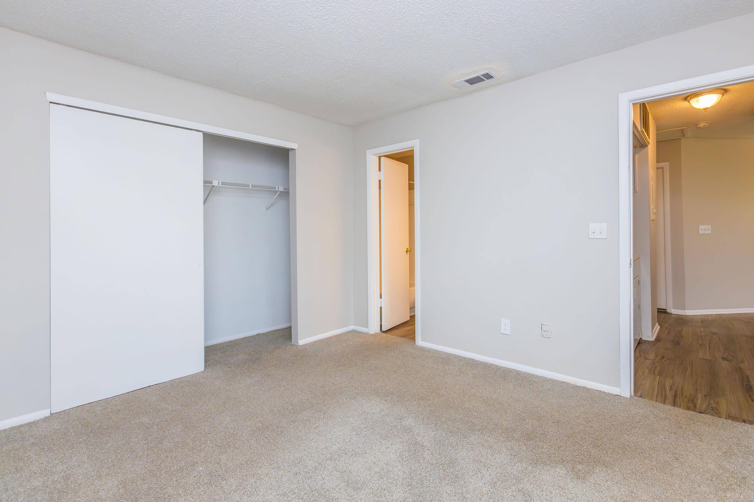 A clean, empty bedroom featuring neutral-colored walls and carpet. There is a large closet with sliding doors on the left and an open doorway leading to another room on the right. Soft lighting enhances the spacious feel of the room, with minimal furnishings.