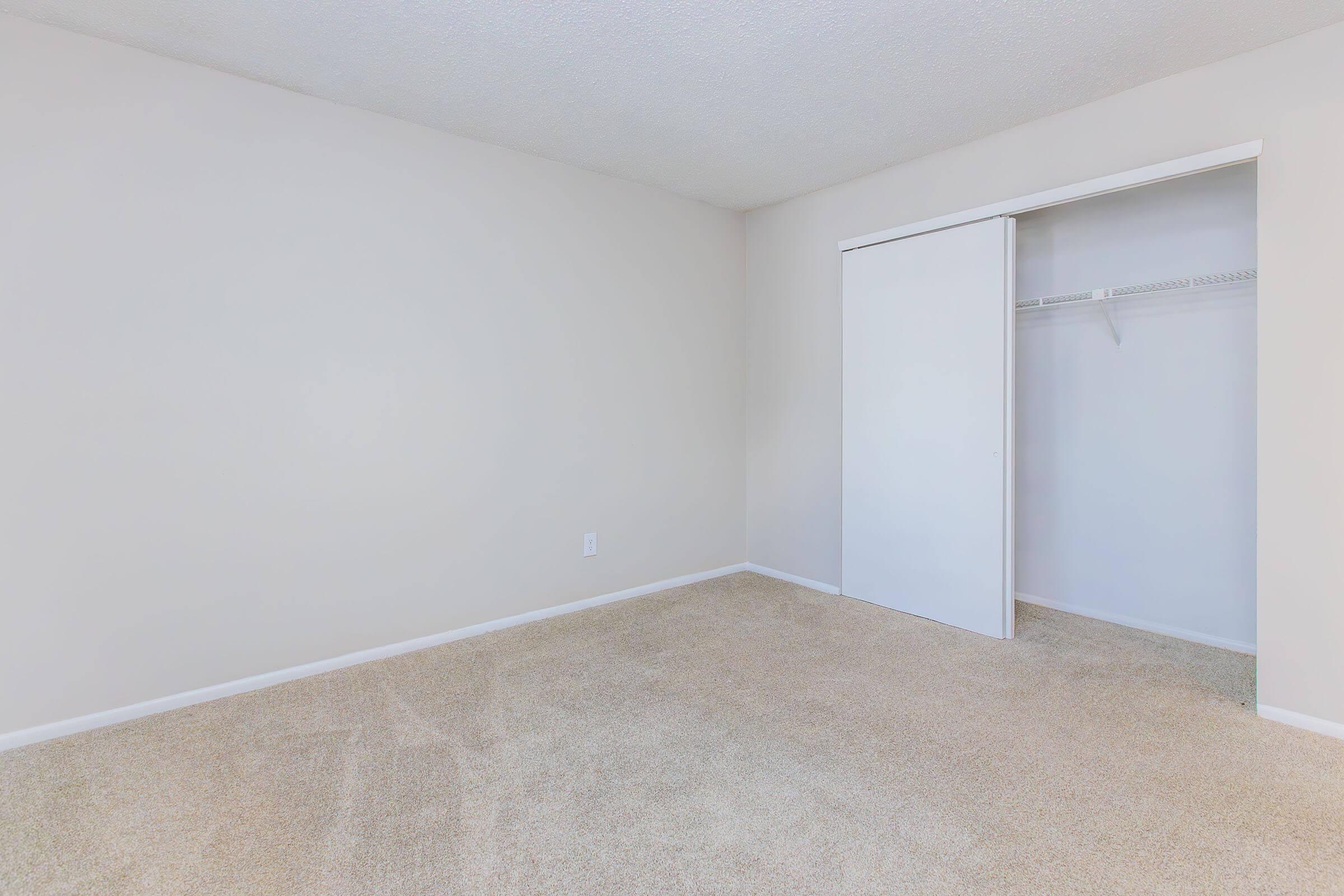 A vacant room with light beige walls and carpet. There’s a white closet with sliding doors on the right side, and the floor is empty, giving a spacious feel. Natural light illuminates the space, making it bright and welcoming. Perfect for customization or moving in furniture.