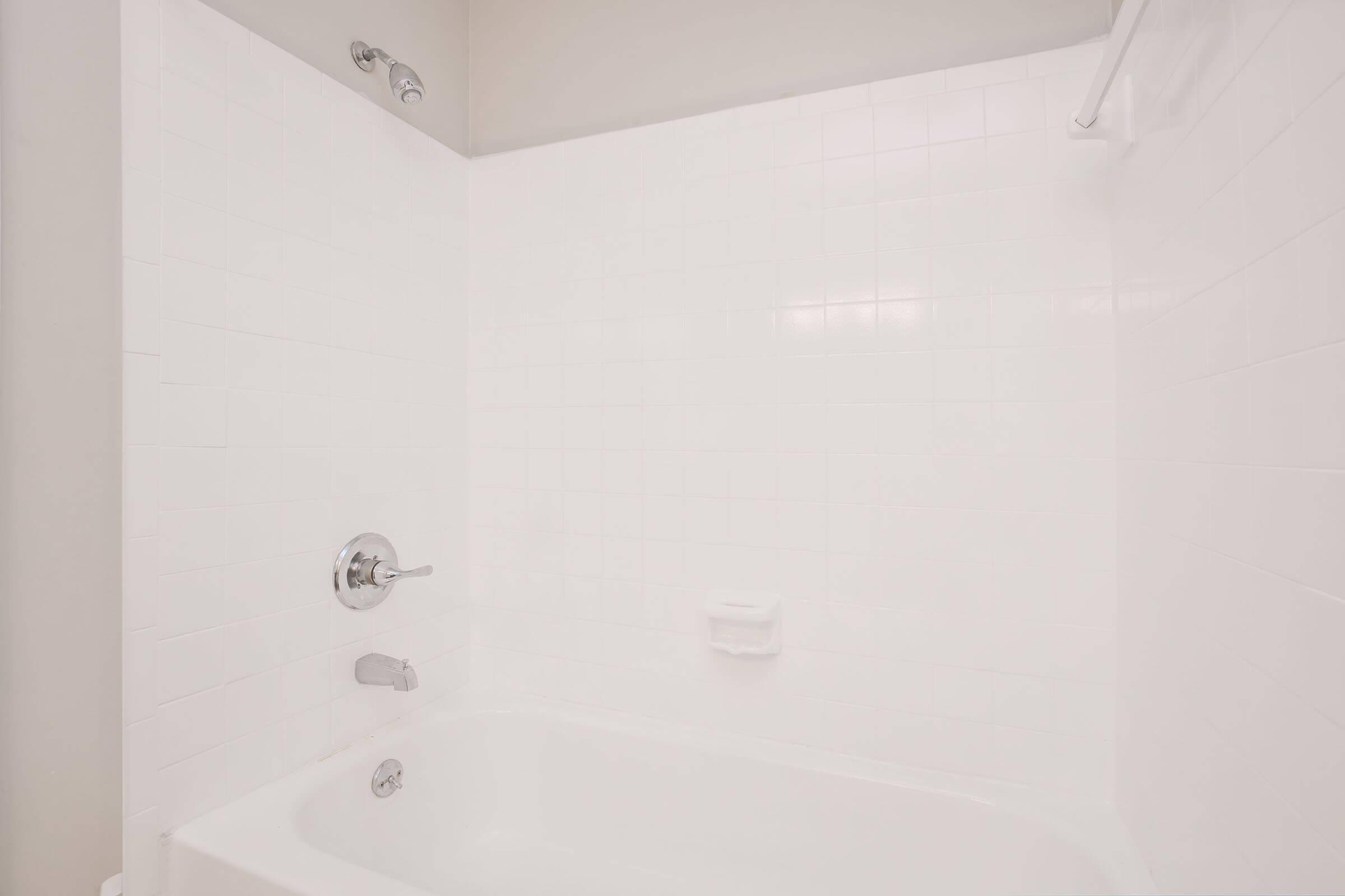 A clean, modern bathtub with a white tiled surround. It features a chrome faucet and showerhead, with a simple soap dish mounted on the wall. The overall appearance is bright and minimalist, emphasizing a spacious, uncluttered bathroom environment.