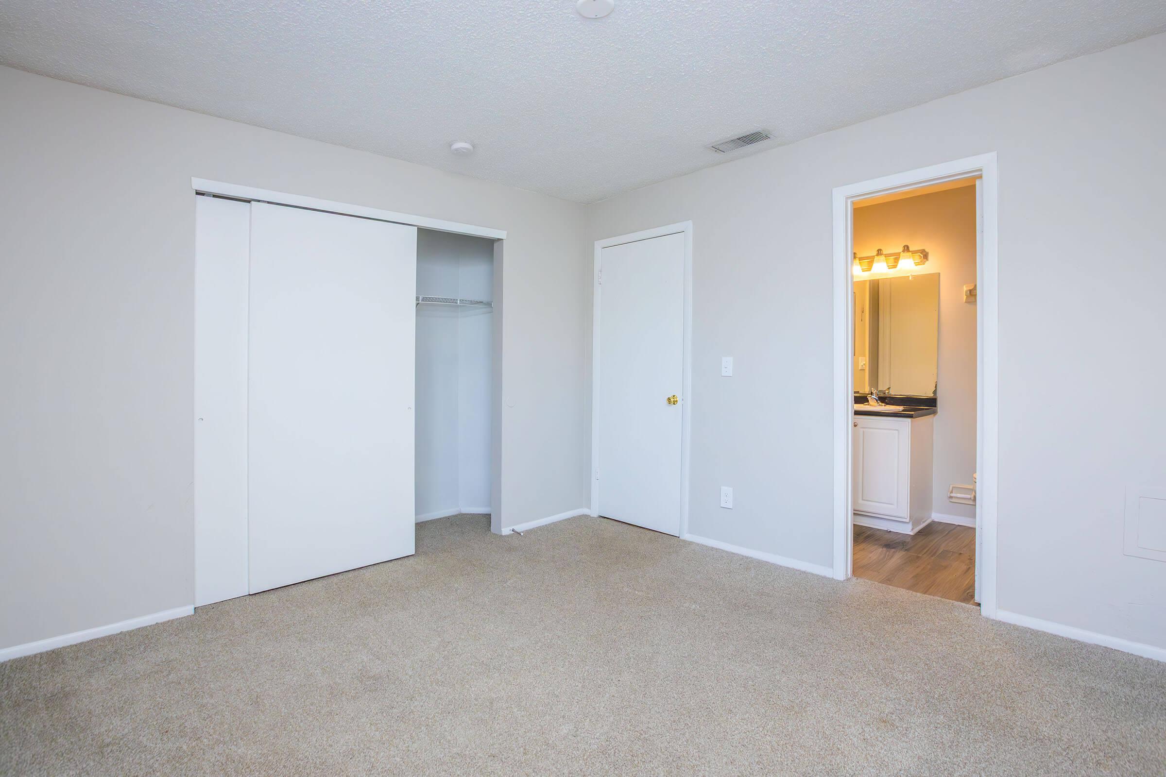 Empty bedroom with light beige carpet and light-colored walls. Features include a closet with sliding doors on the left, a closed door leading to a bathroom on the right, and a plain ceiling. The room has a bright, airy atmosphere with minimal furnishings.