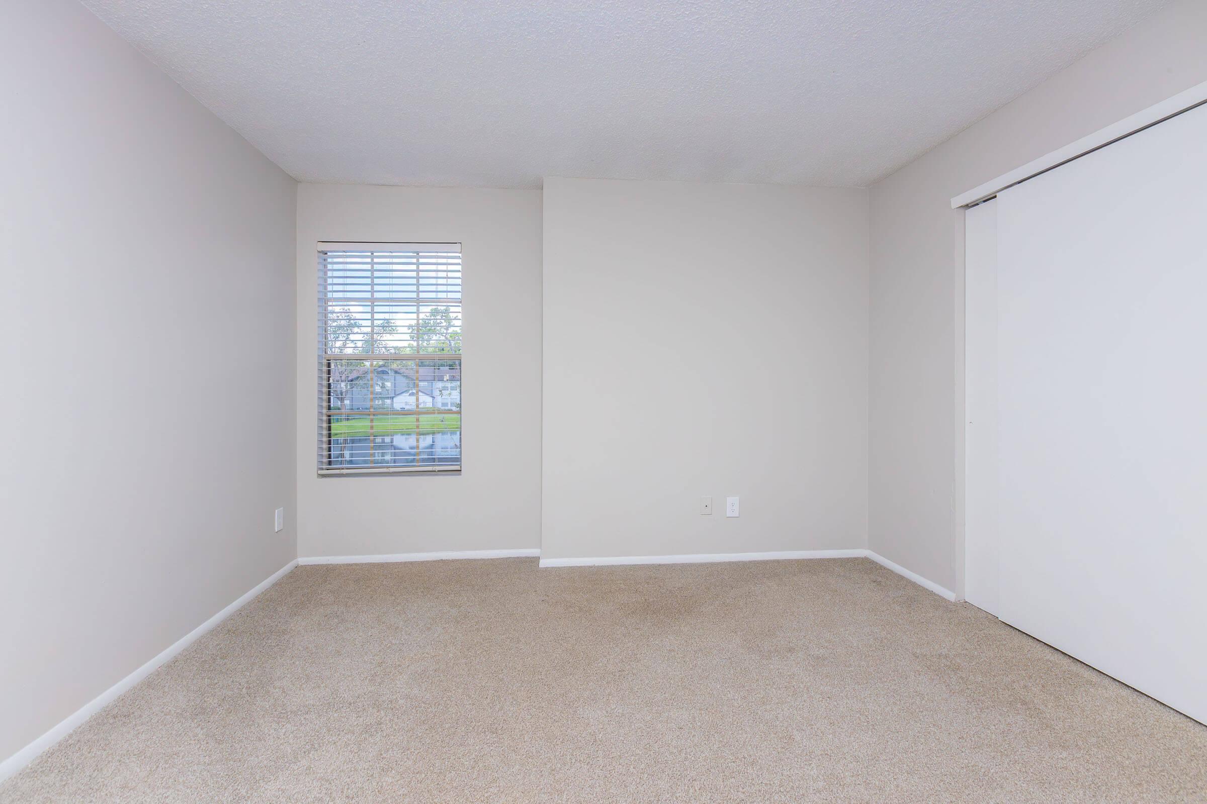 A sparse room with beige carpet and light gray walls. There is a window with white blinds on the left side, allowing natural light to enter. A closet door is on the right side, and the overall design is minimal, creating a clean and open space.