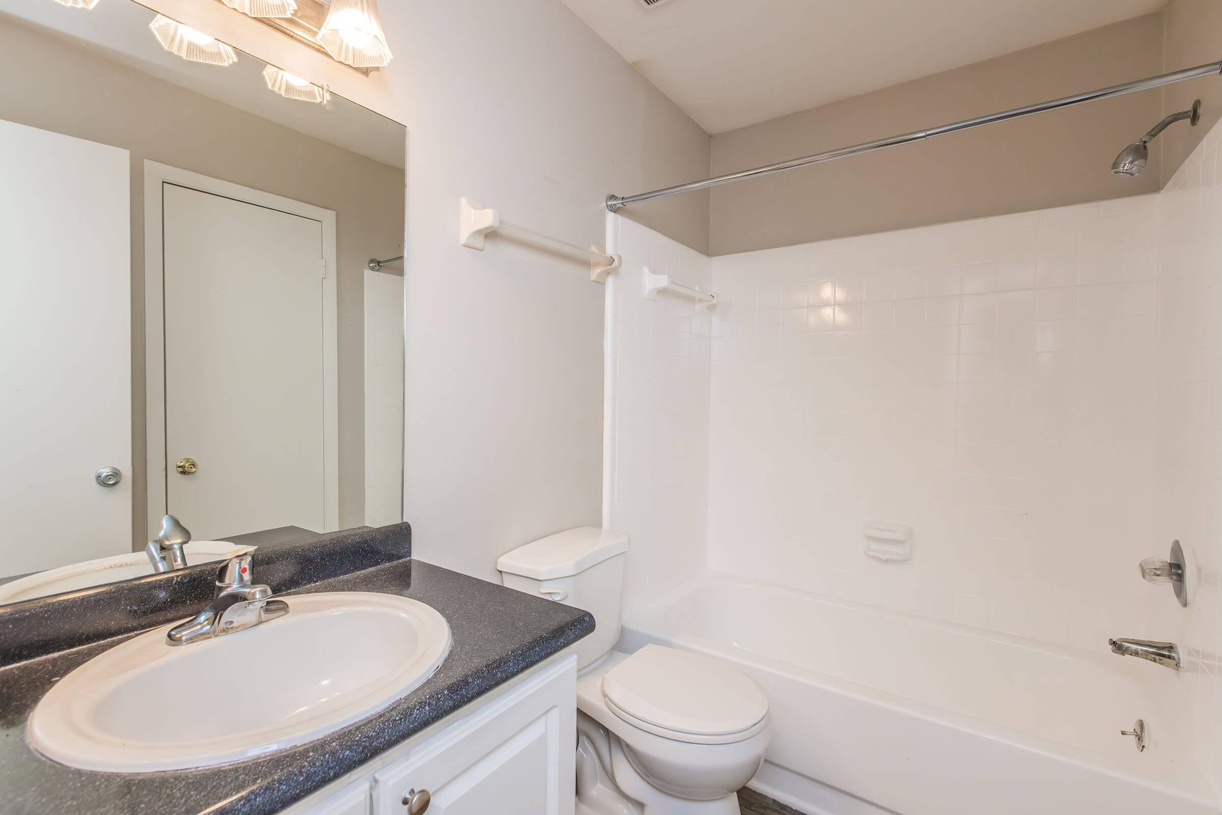 A clean and simple bathroom featuring a white bathtub with a shower, a sink with a dark countertop, and a toilet. The walls are light and there is a mirror above the sink. A light fixture with multiple bulbs is visible above. A door leads to another room, providing a minimalist aesthetic.
