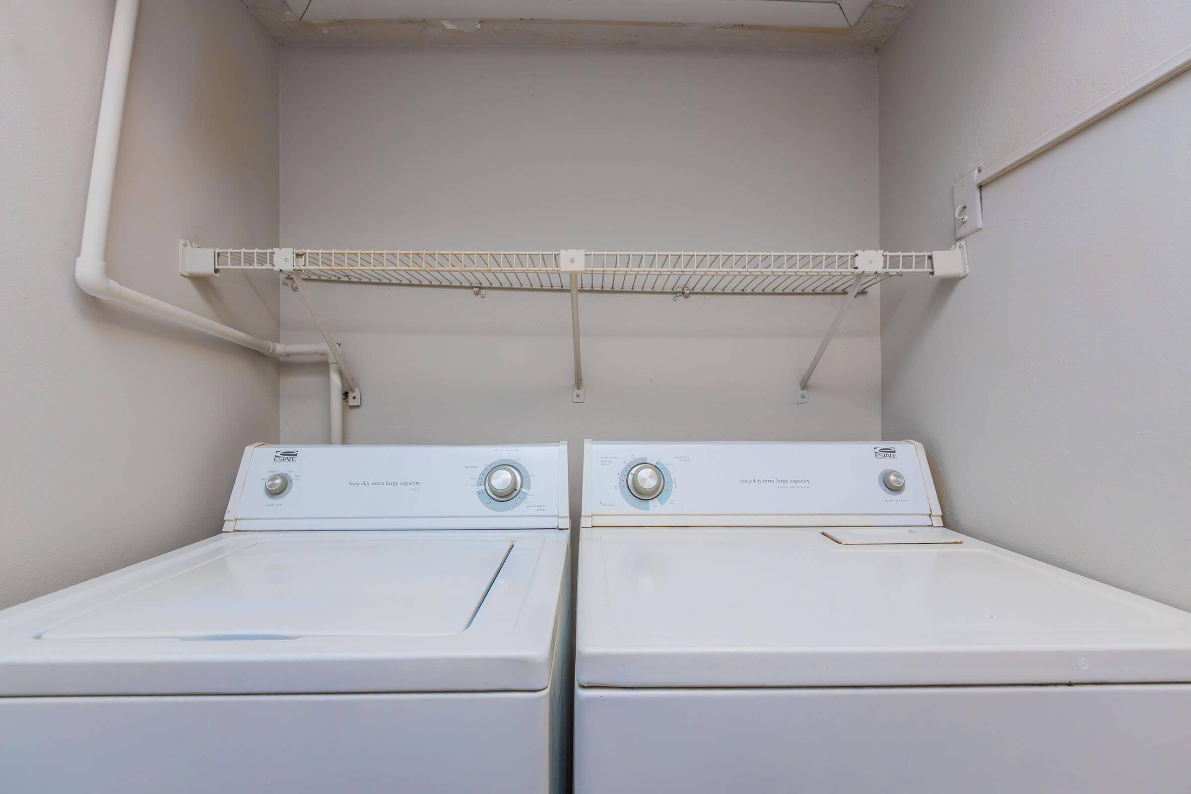 A clean laundry room featuring a stacked washer and dryer on the left side. Above the appliances is a metal shelf for storage, with plain white walls surrounding the area, creating a functional and organized space for household laundry tasks.